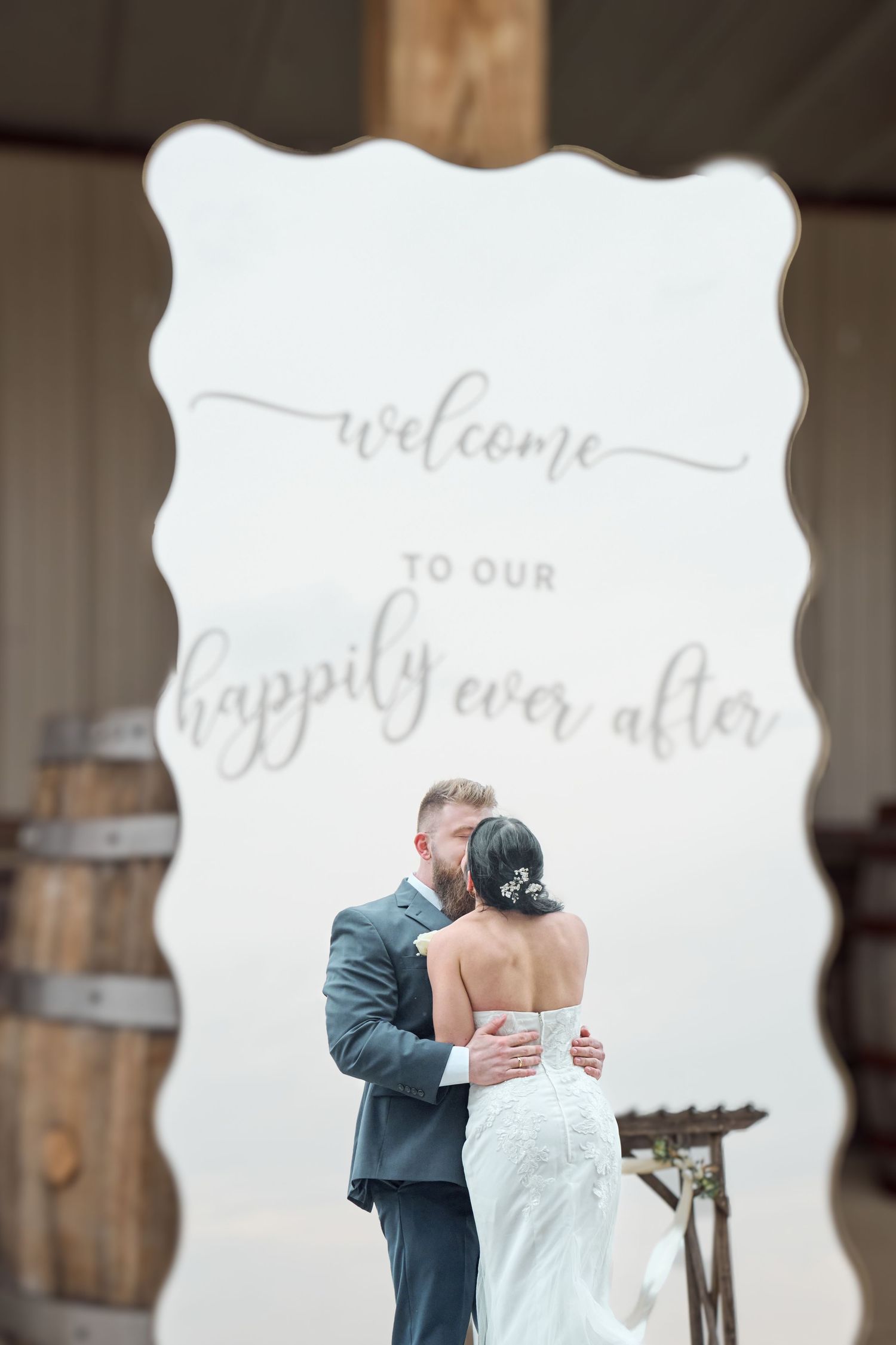 Couple embraces under 'Welcome to our happily ever after' sign at rustic wedding venue.