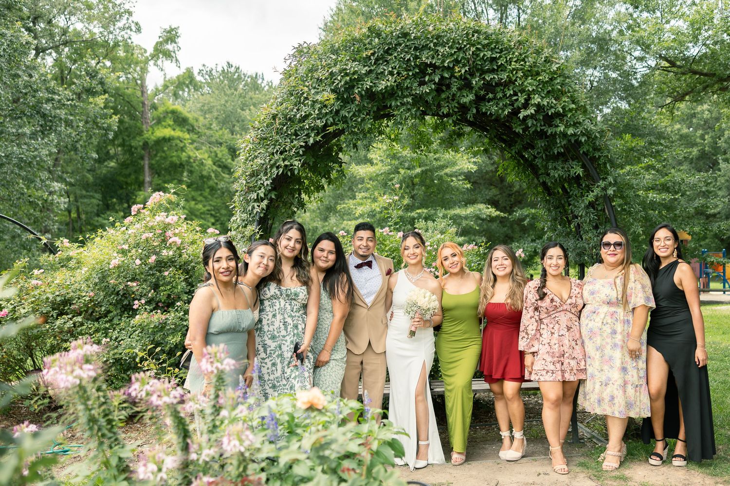 Group photo of wedding guests standing together under a lush garden archway with flowering plants.