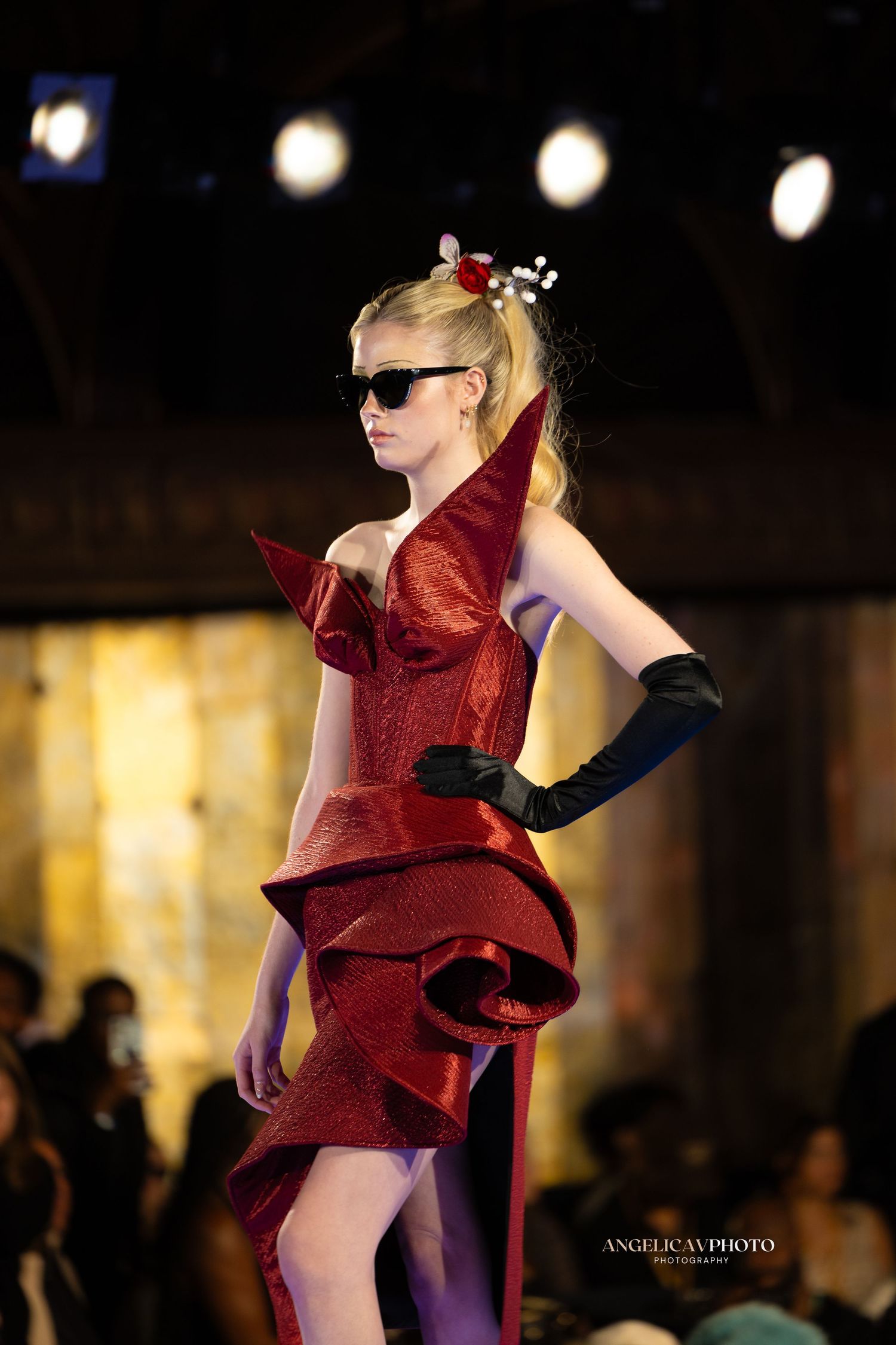 Model wearing red sculptural cocktail dress with black gloves and sunglasses on catwalk.