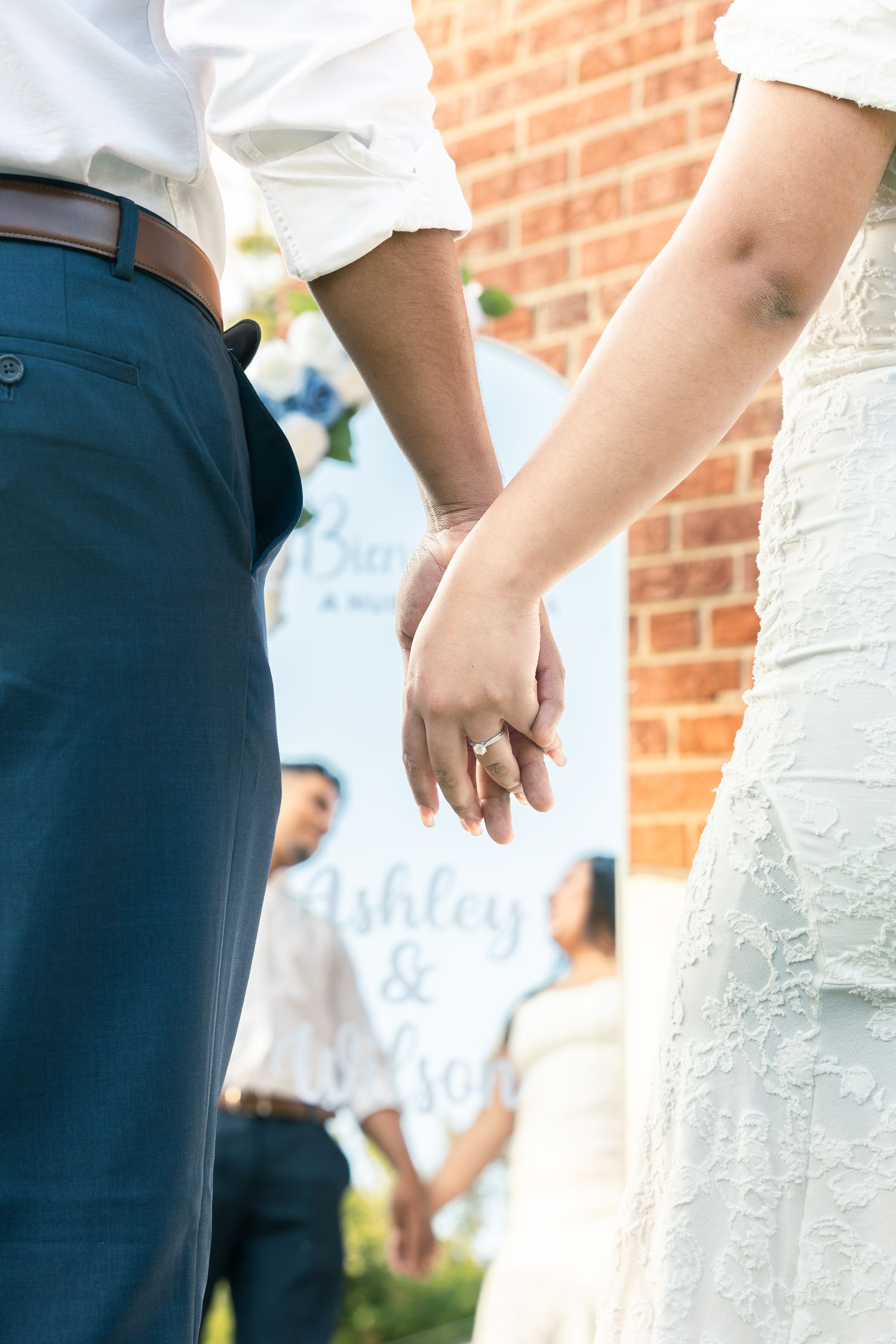 Close-up of a couple holding hands at a wedding with a brick wall background.
