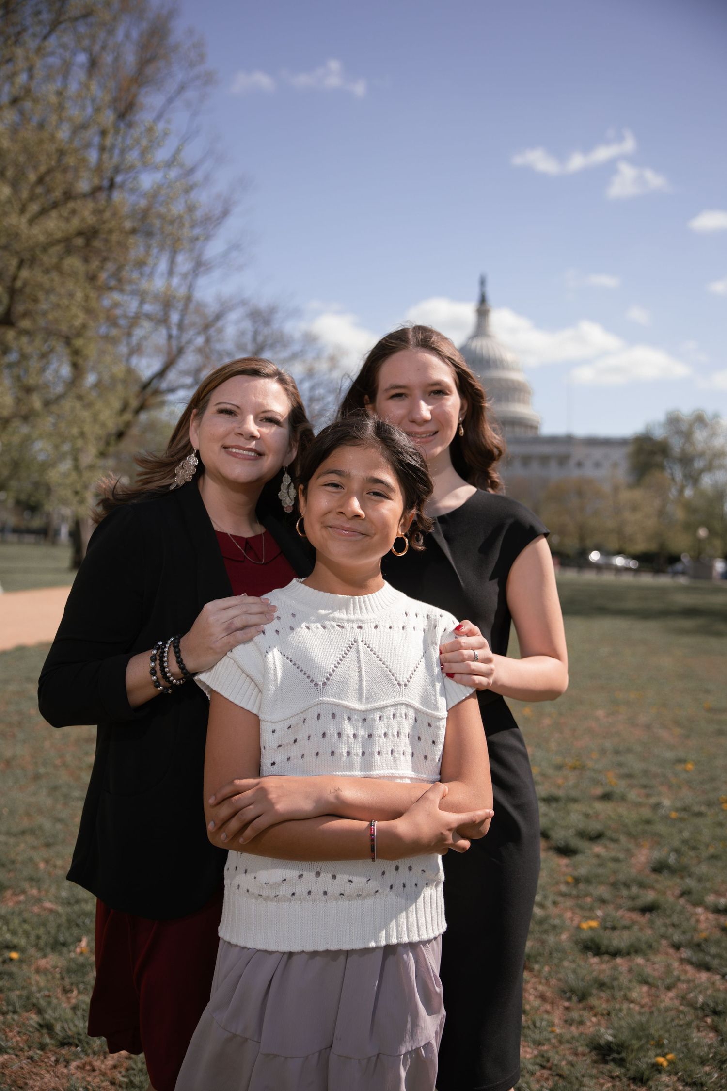 Three people pose together for a photo with the United States Capitol dome visible in the background on a sunny day.