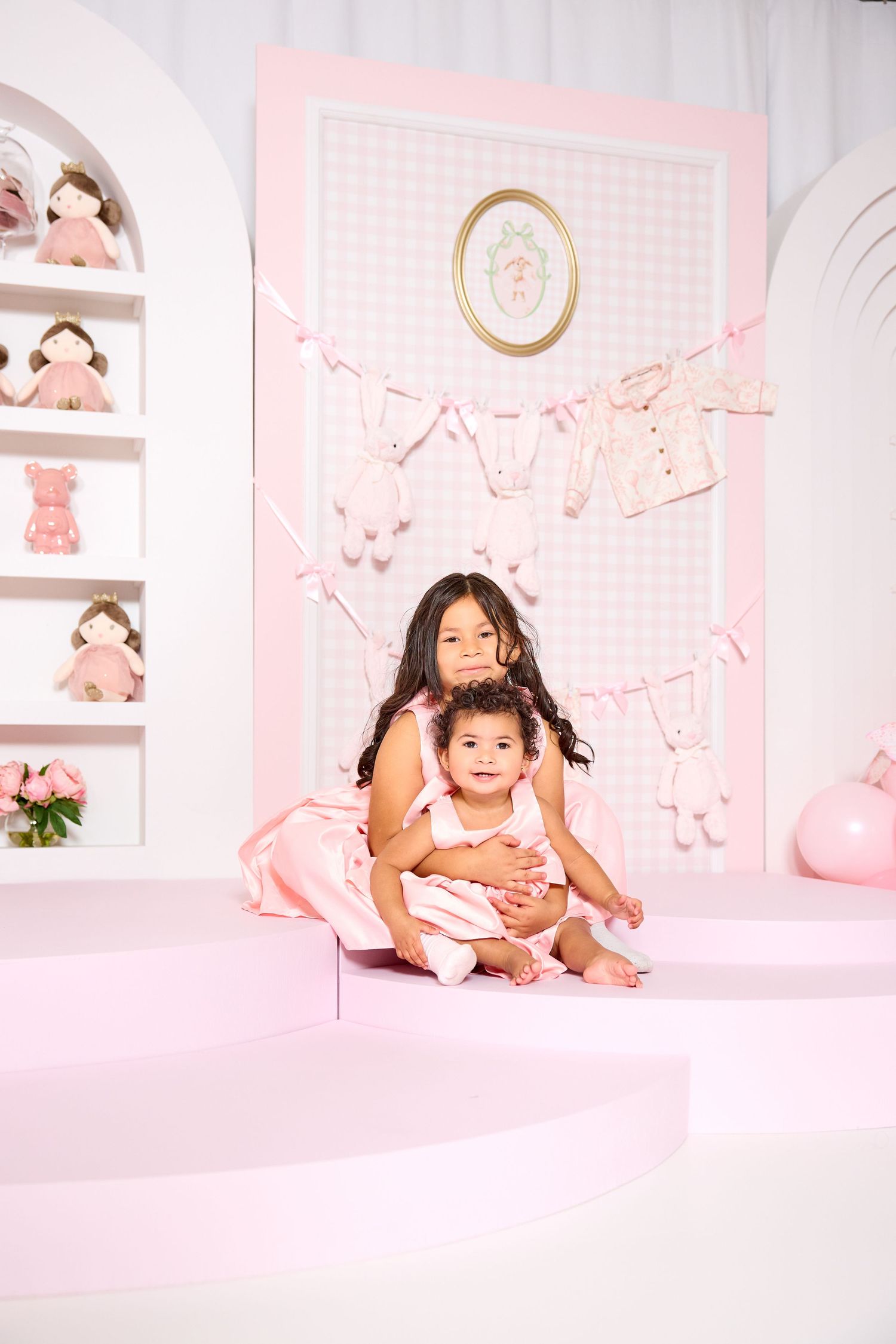 Two young sisters in pink outfits pose together at a baby-themed photo shoot with dolls and pastel decorations.