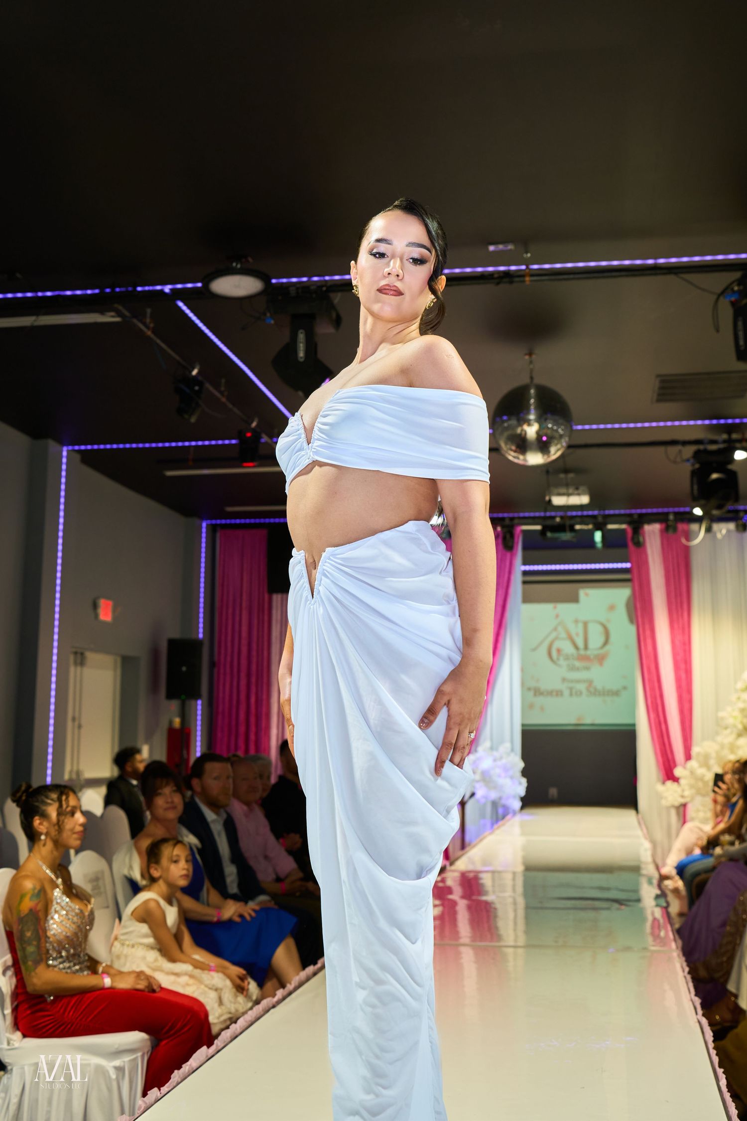 Model walks runway in light blue off-shoulder crop top and draped skirt at fashion show.