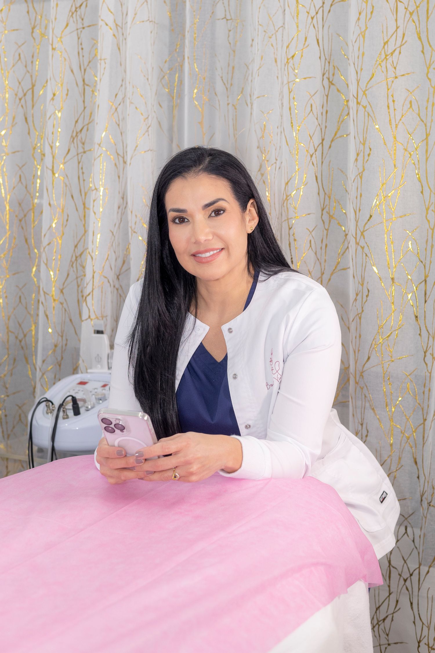 Healthcare professional in white cardigan and navy top sits at pink treatment table while checking mobile phone.