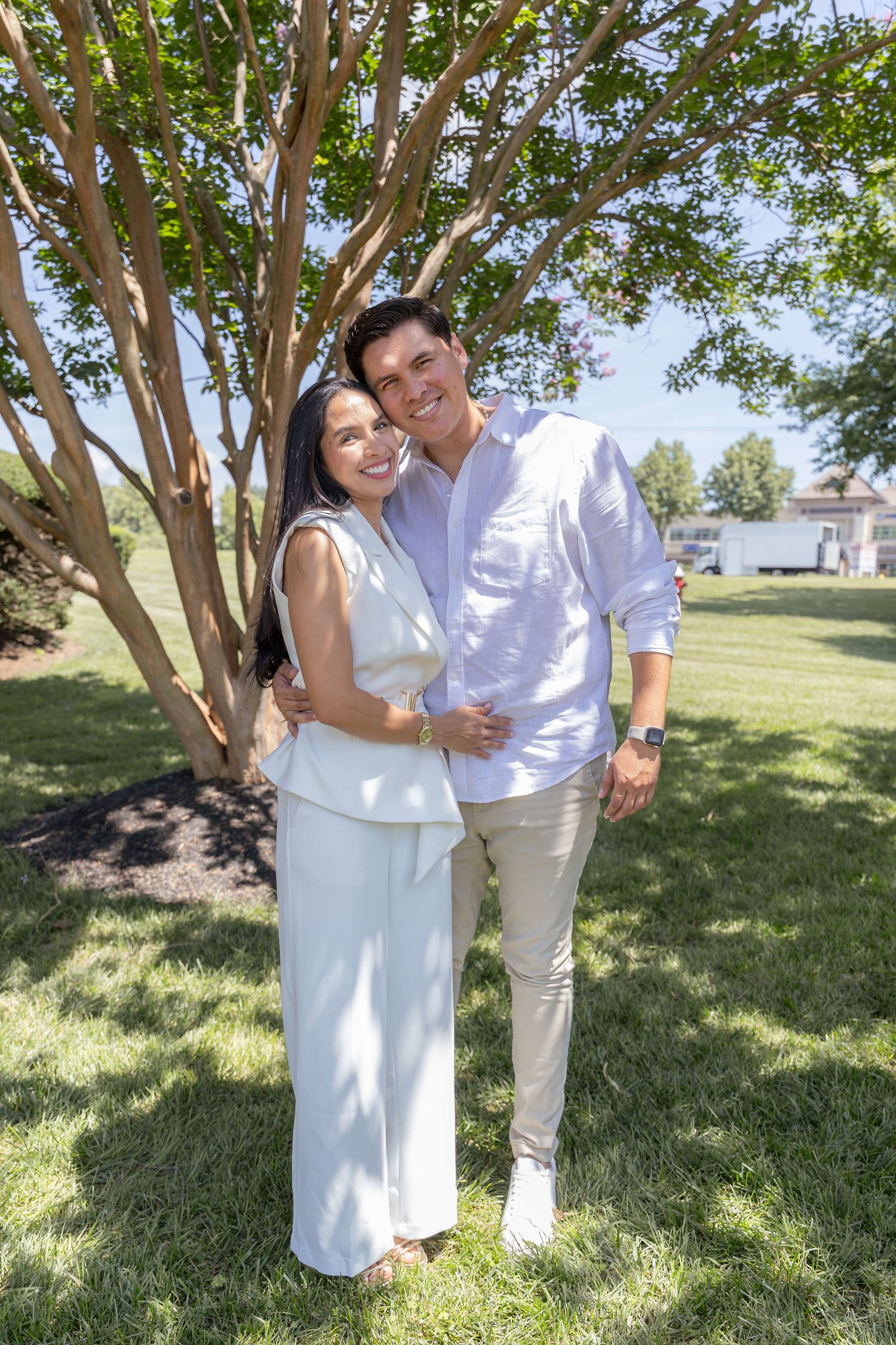 A couple shares a tender moment while standing together in white outfits under a large tree.