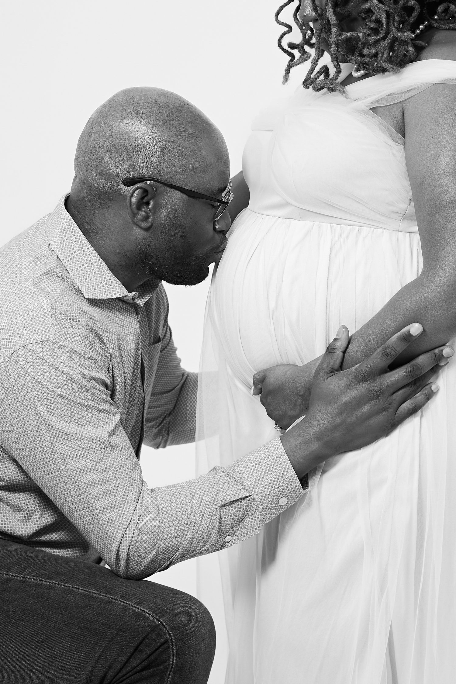 Black and white maternity photo of a man kissing pregnant woman's belly while kneeling beside her.