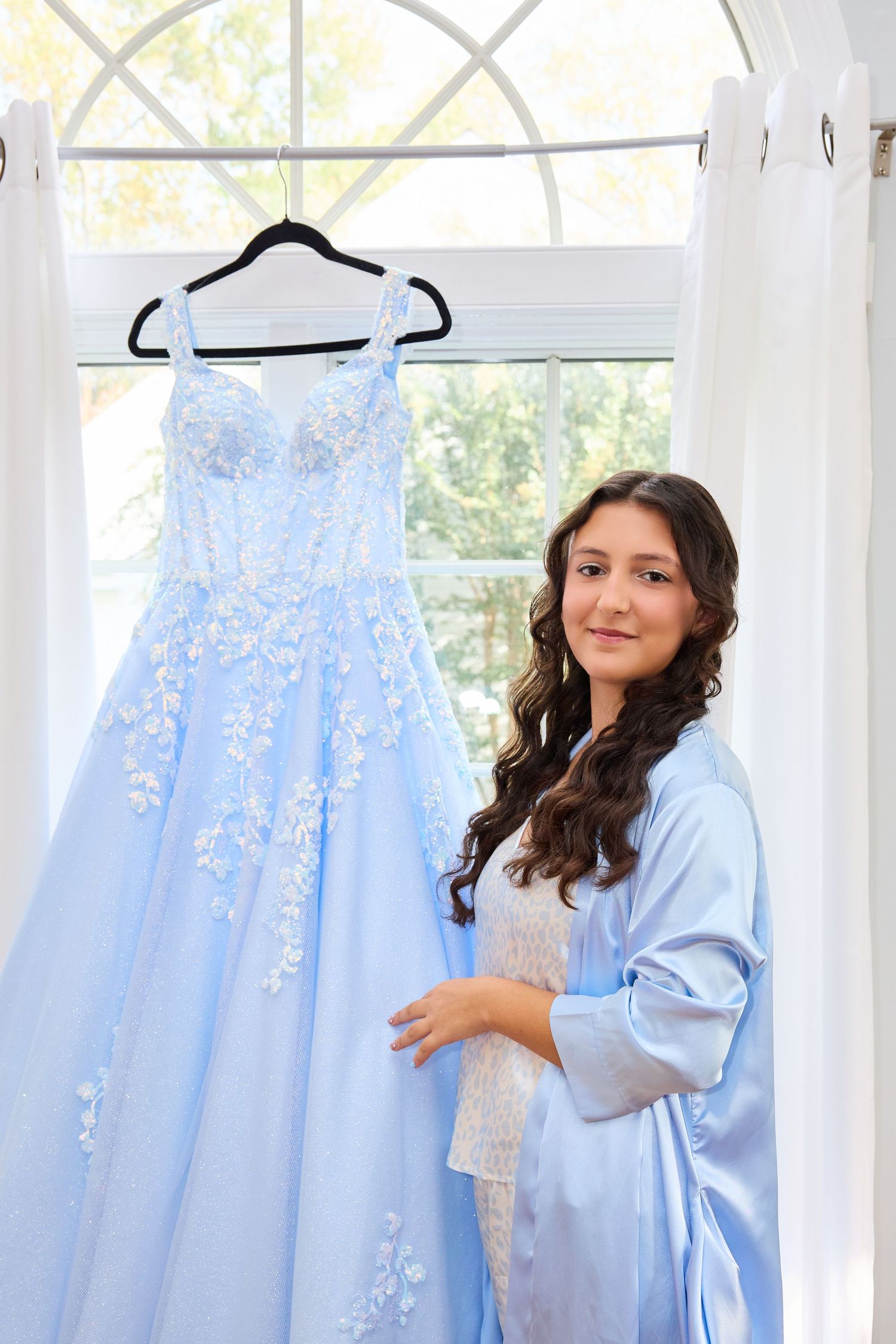 Light blue bridesmaid dresses hang on display in a sunlit room with white curtains.