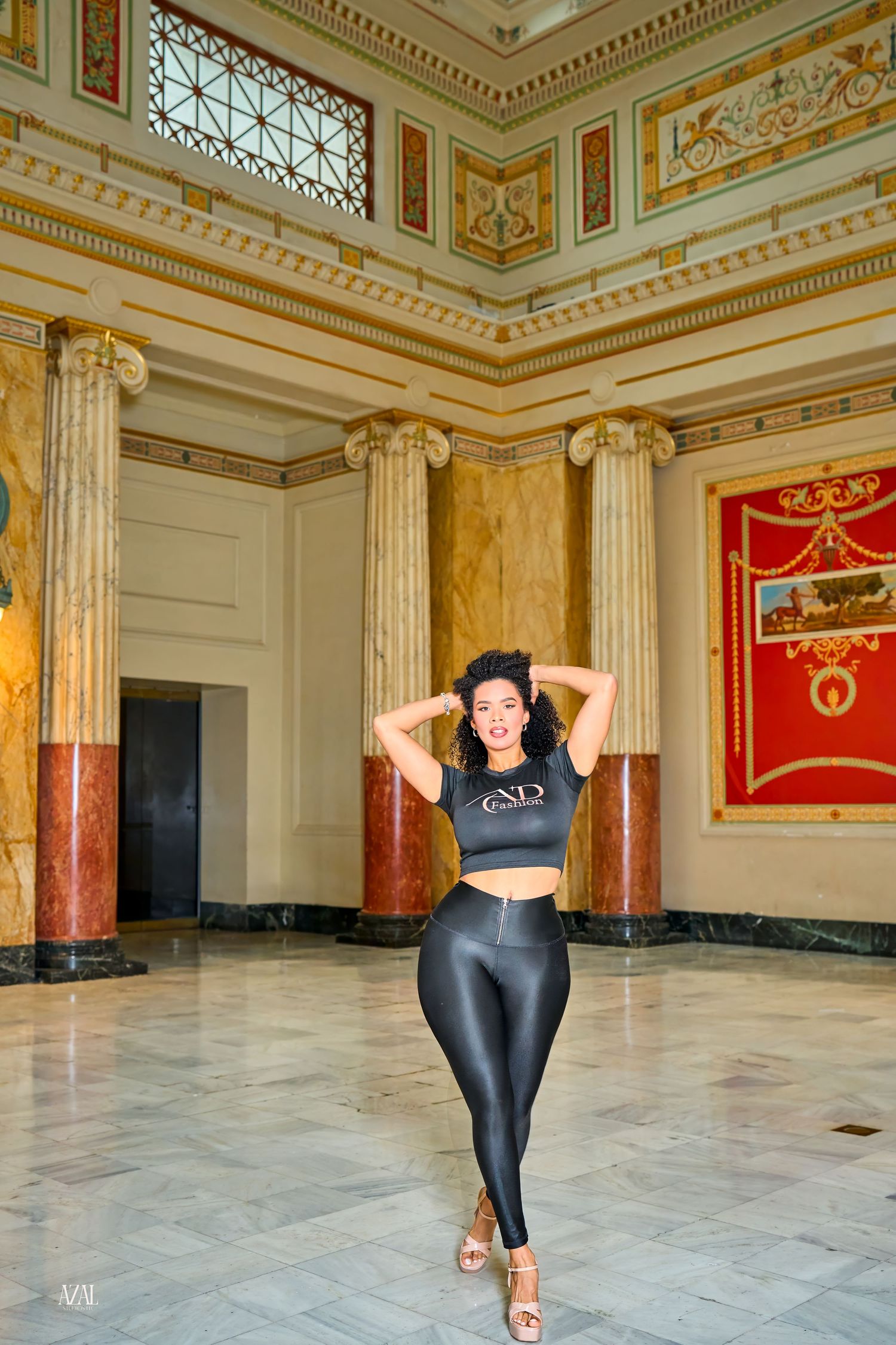 Woman in black crop top and shiny leggings poses confidently in ornate classical building with marble columns.