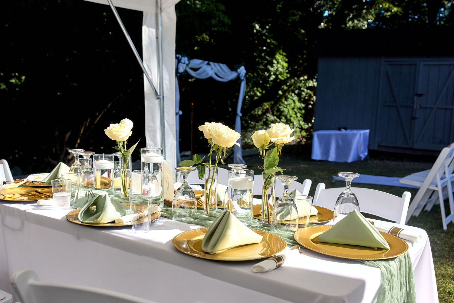 An elegant outdoor wedding table setting with gold chargers, pale yellow napkins and white roses as centerpieces.