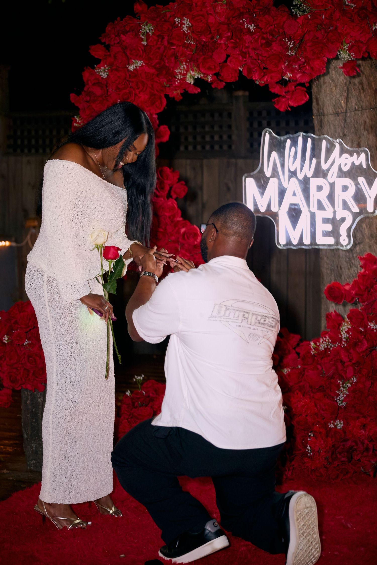 A romantic marriage proposal setup with illuminated letters spelling 'WILL YOU MARRY ME' against a backdrop of red roses.