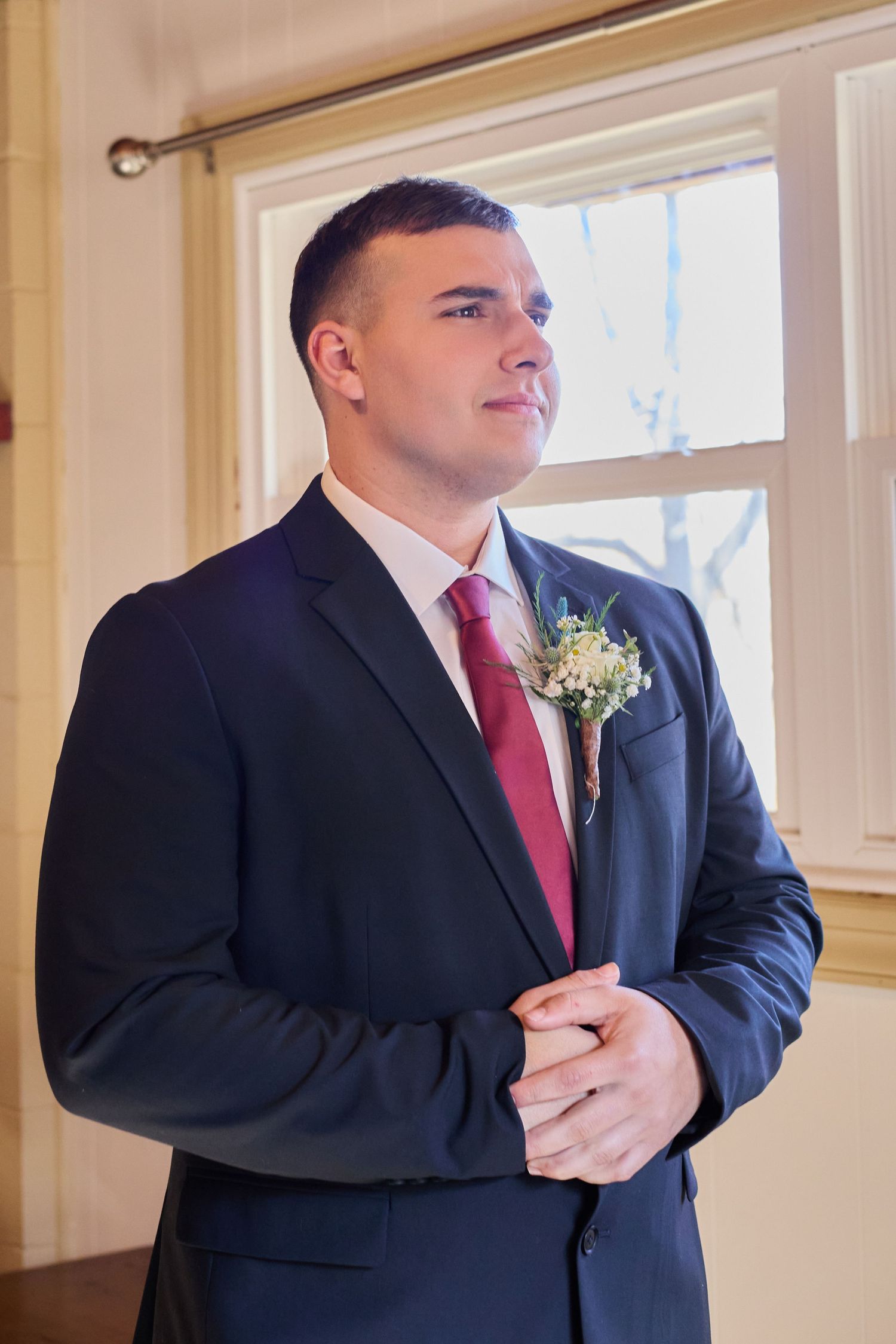 A groom in a navy suit with red tie and boutonniere stands by a window during a wedding ceremony.
