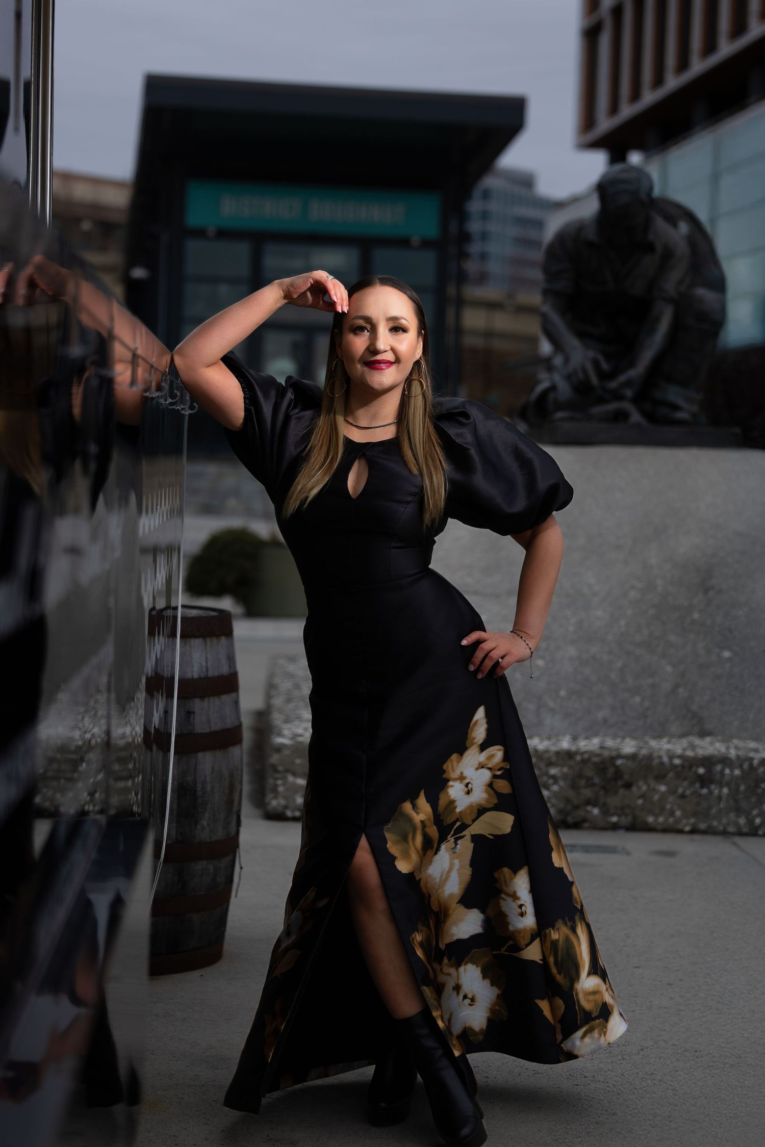 Dramatic fashion pose showcasing black evening gown with floral pattern against urban backdrop.