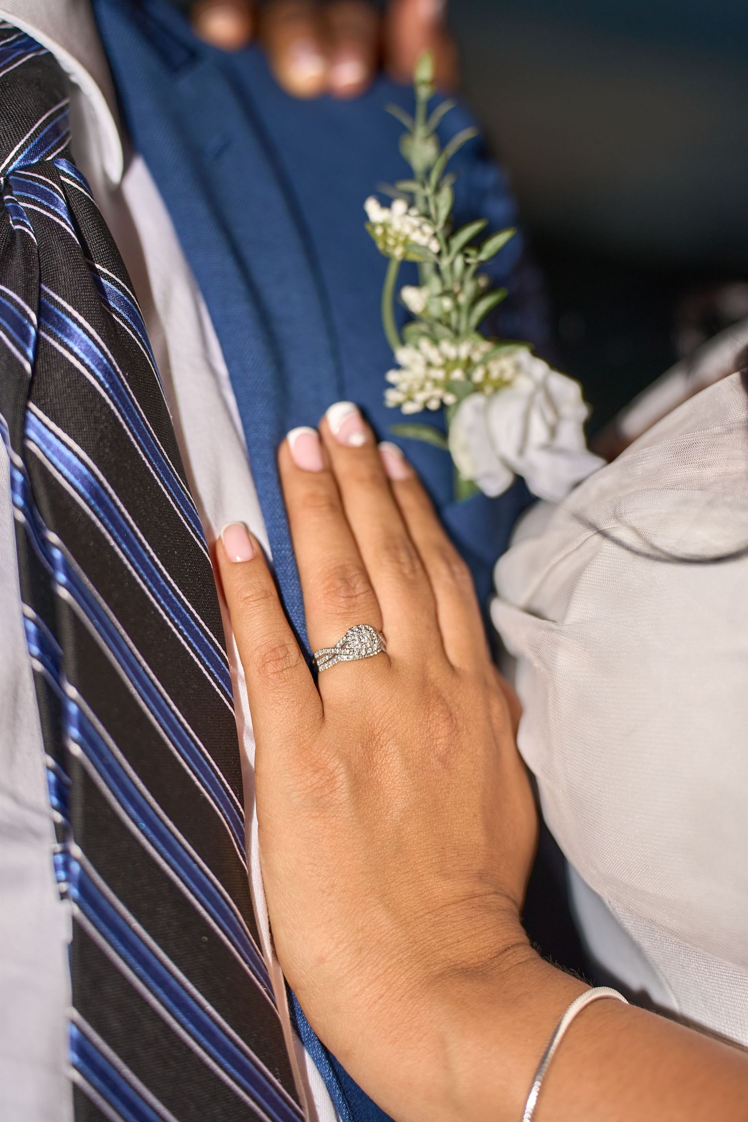 Close-up of a bride's hand with a diamond engagement ring resting on groom's suit with striped tie and boutonniere.