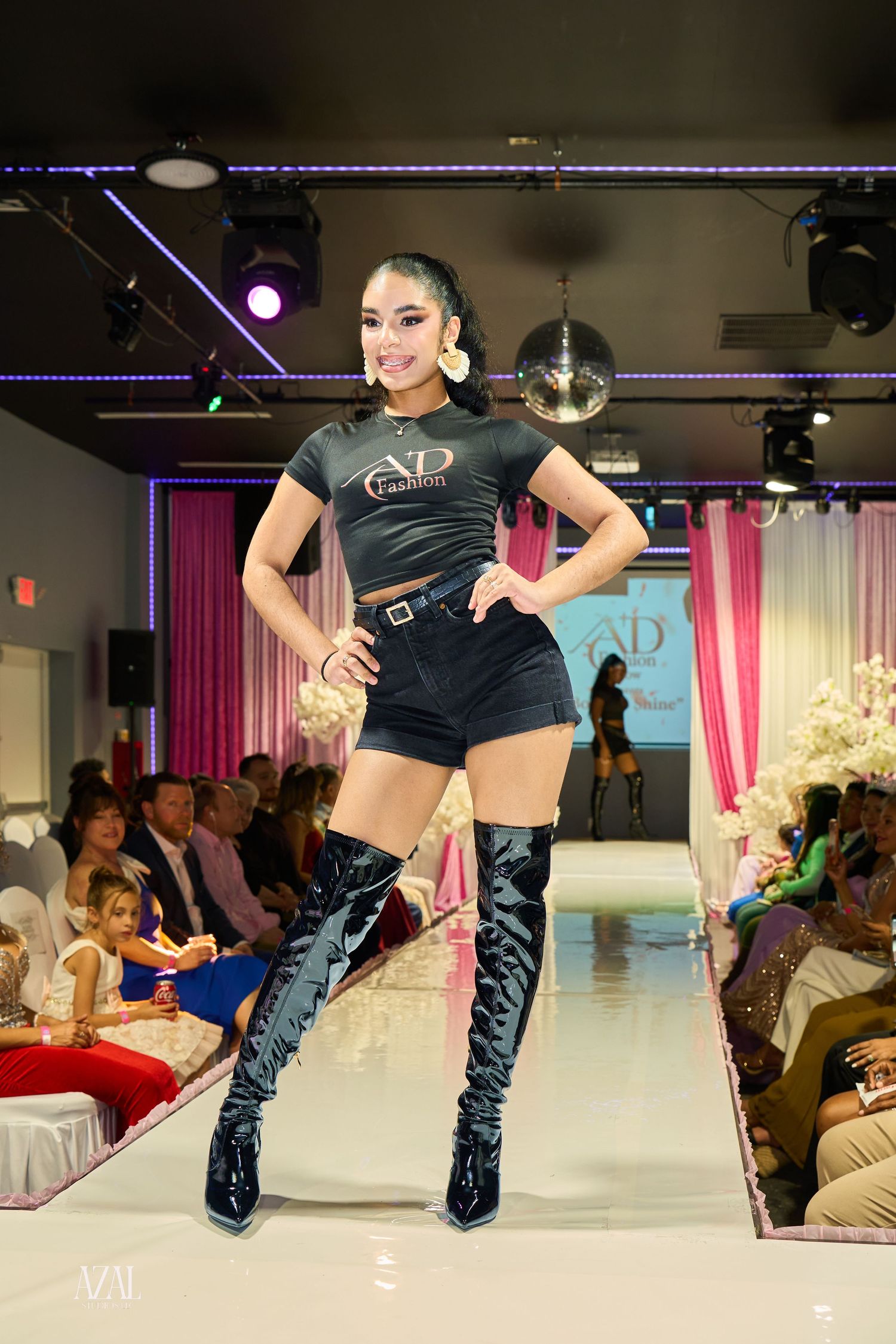 Model wearing AD Fashion black tee, shorts, and thigh-high boots walks the runway at a fashion show.