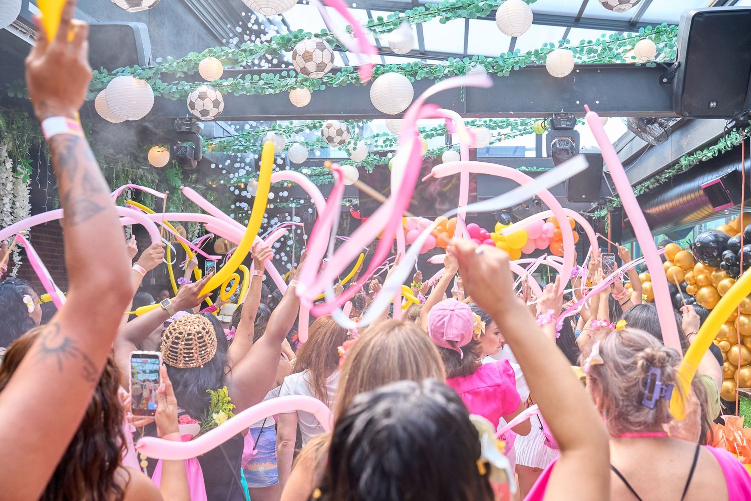 Crowd of party guests waving pink streamers and yellow ribbons under disco balls at a vibrant indoor celebration.