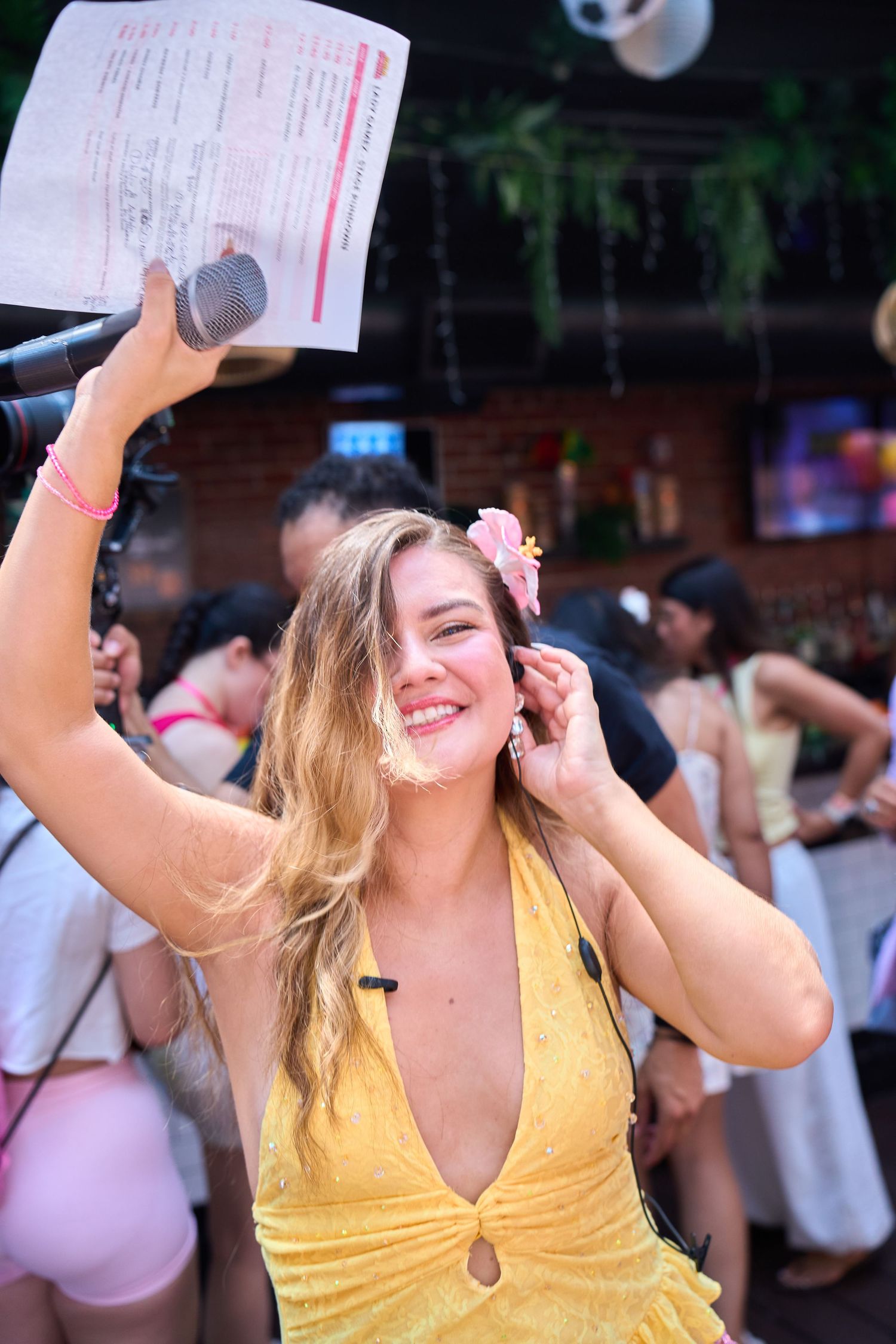 Smiling blonde woman in yellow dress holding microphone and paper at a lively party event.