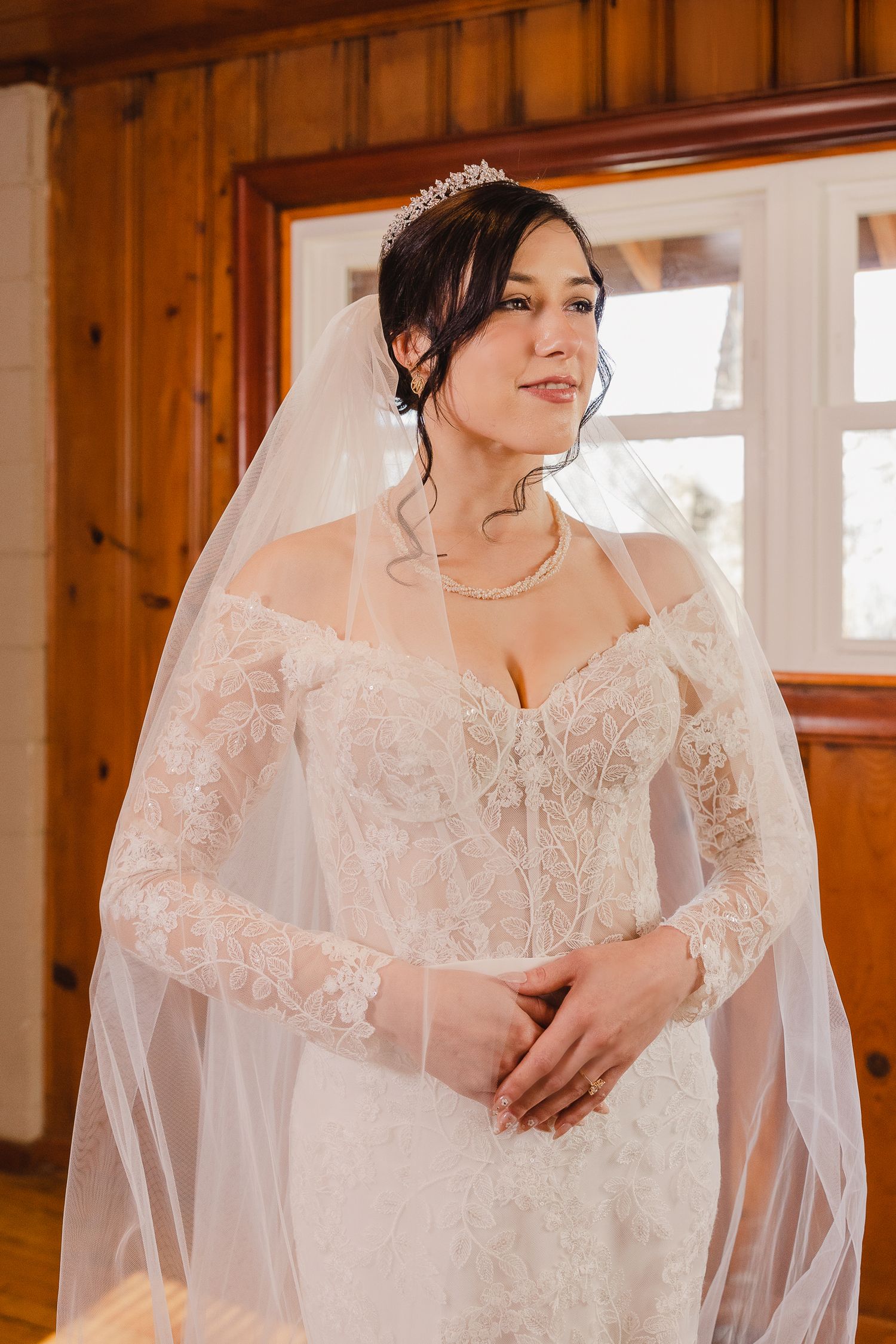 A bride in a detailed lace wedding dress and veil poses in a rustic wooden room with natural lighting.