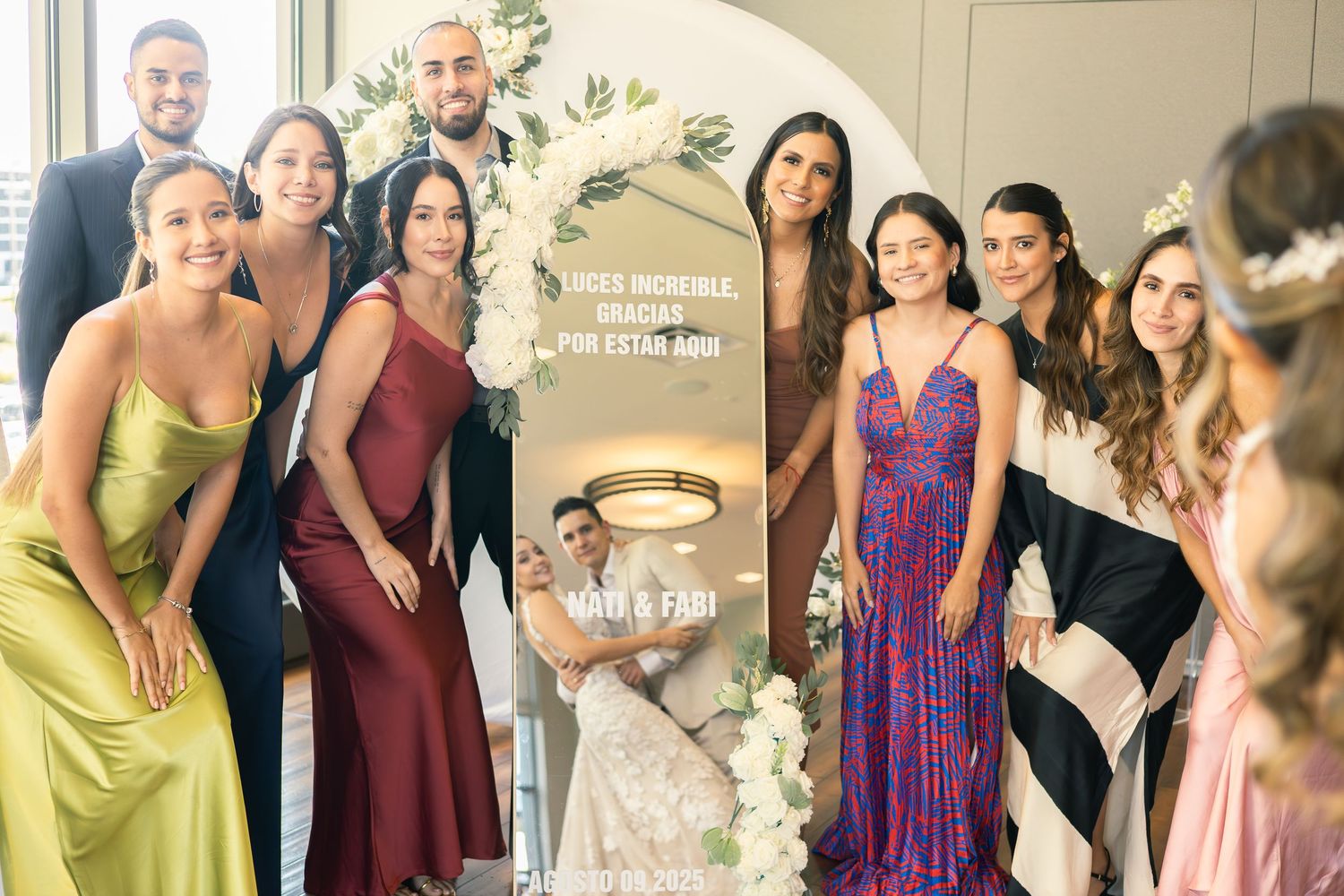 A wedding party in formal attire poses together in front of a mirror at an elegant venue.