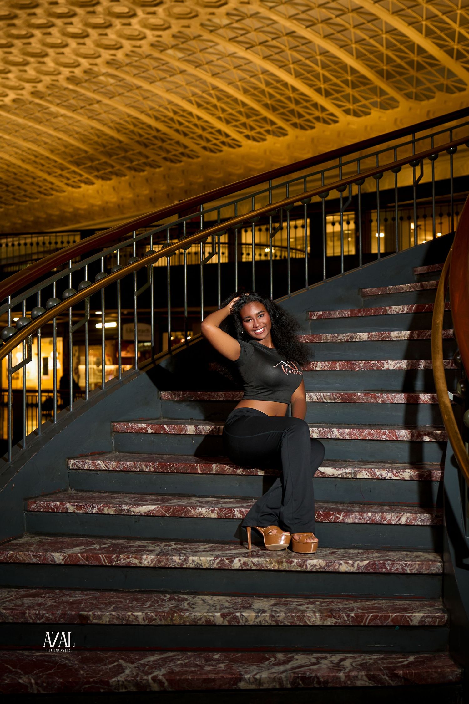 Woman in black crop top and pants poses gracefully on ornate marble staircase with golden ceiling in elegant venue.