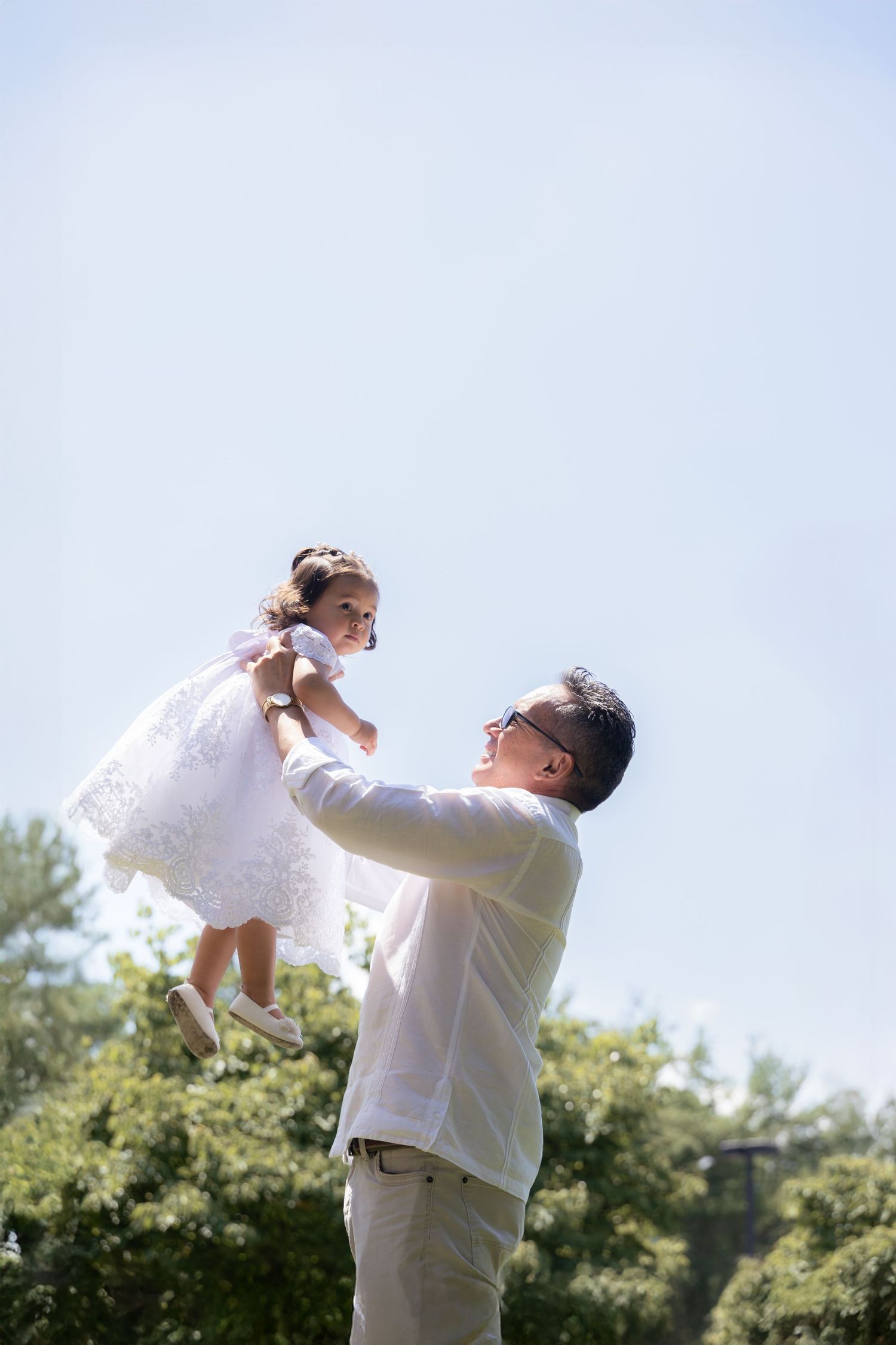 Person in white lifting child wearing lace dress against blue sky background.