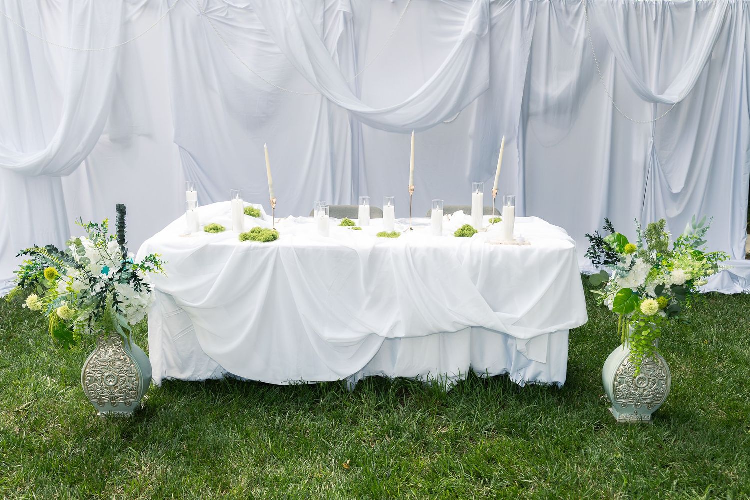 Wedding reception setup with white tablecloths and chairs on a green lawn under a white tent canopy.