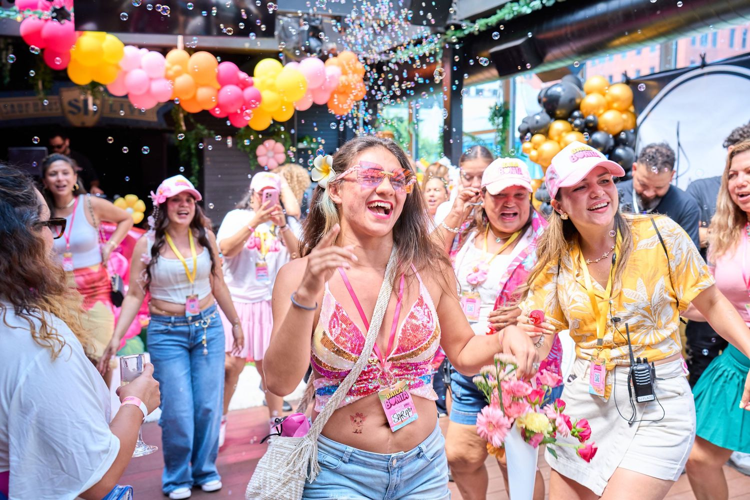 Colorful festival crowd of young women celebrating with powder paint, balloons, and confetti outdoors.