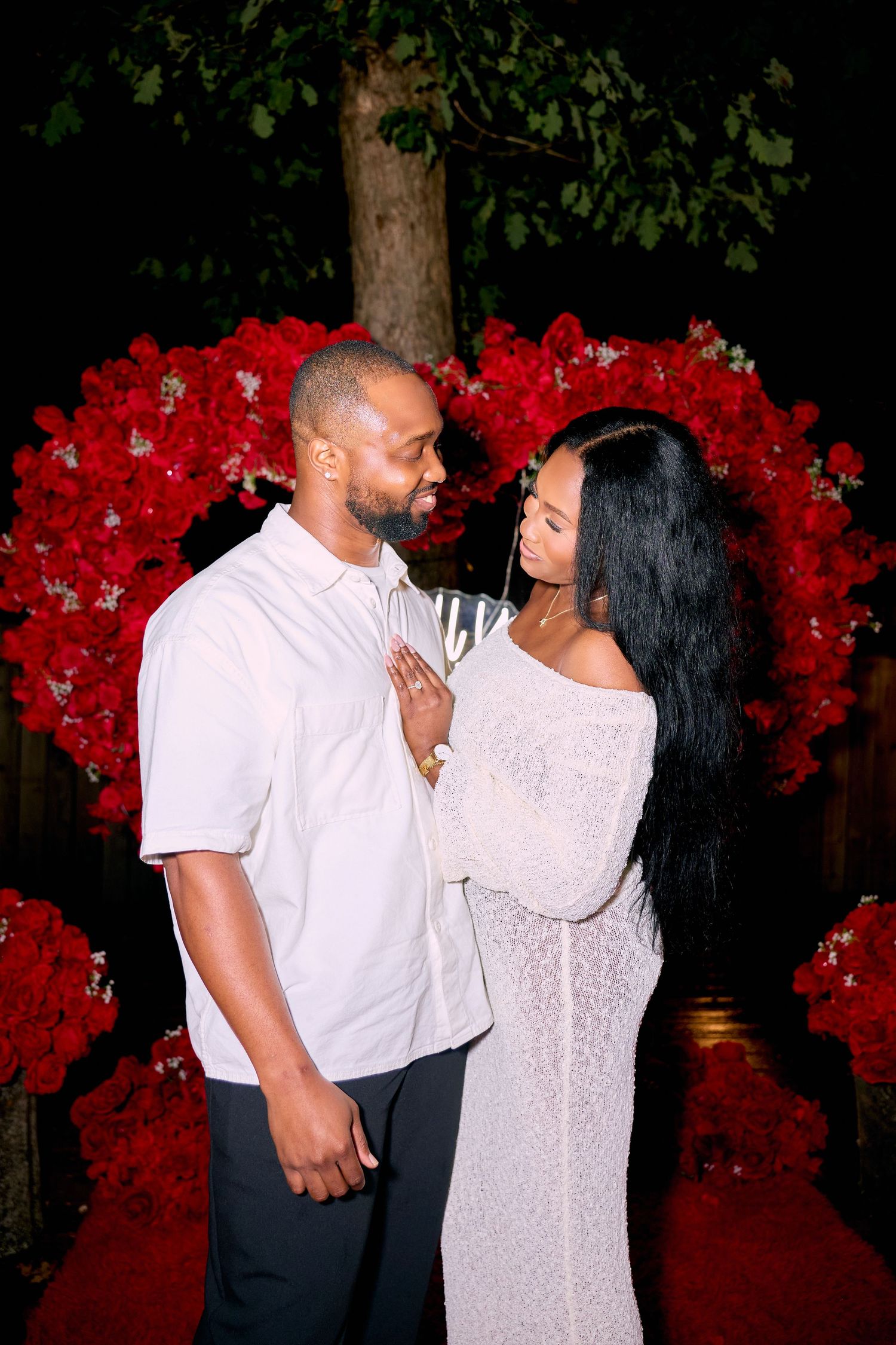 A couple sharing intimate moments in front of a romantic red floral backdrop with glowing candles.