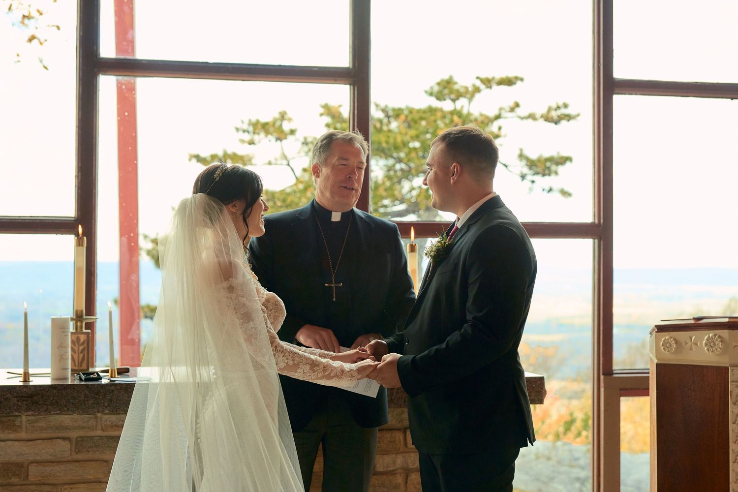 A wedding ceremony takes place in front of large windows with scenic mountain views in the background.