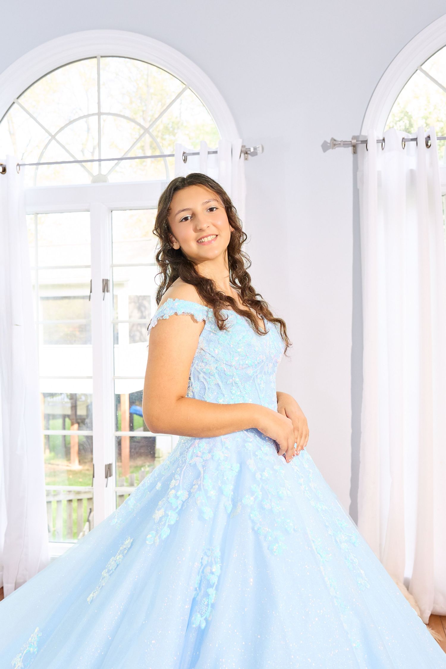 A young person wearing a light blue ball gown twirls in an elegant white room with arched windows.