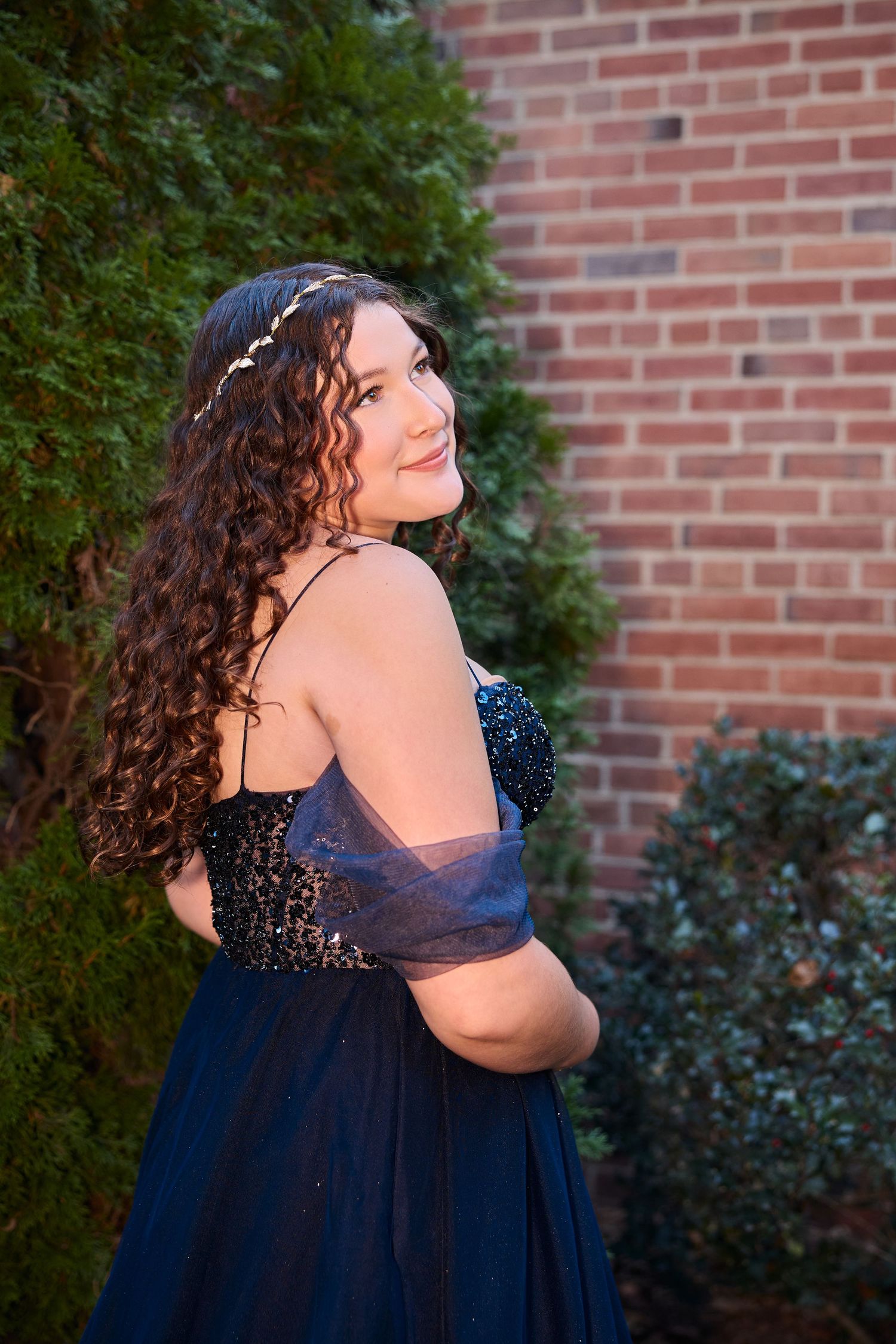A person in a navy blue formal dress with curly hair poses against a brick wall background.