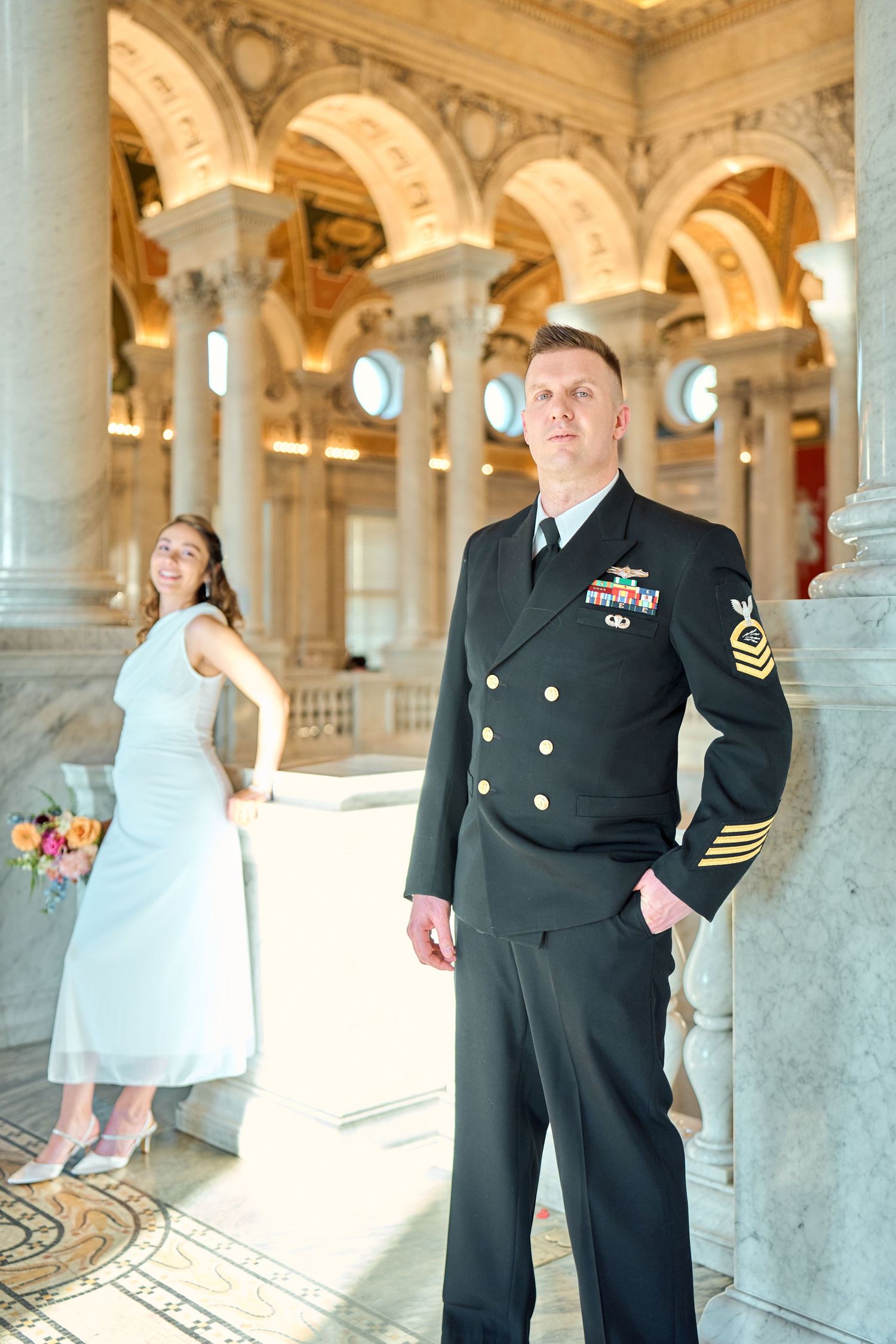 Navy officer in dress uniform stands beside a bride in white gown inside an ornate marble building.