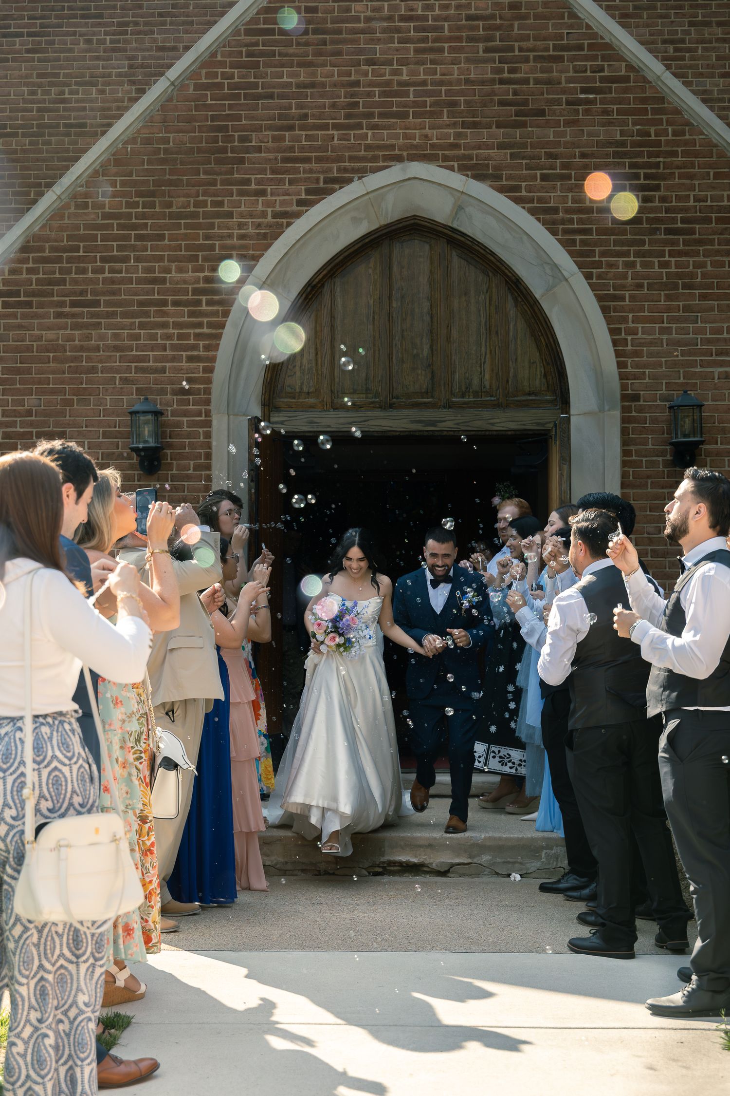 Newlyweds exit church while guests blow bubbles and celebrate under gothic brick archway entrance.