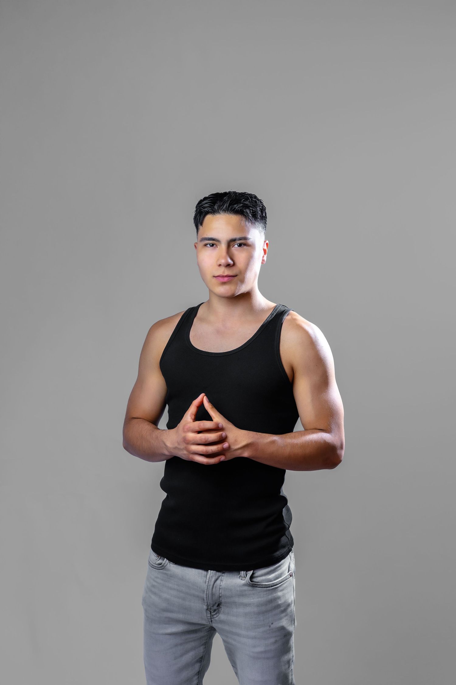Athletic person in a black tank top and grey jeans poses confidently with clasped hands against a grey background.