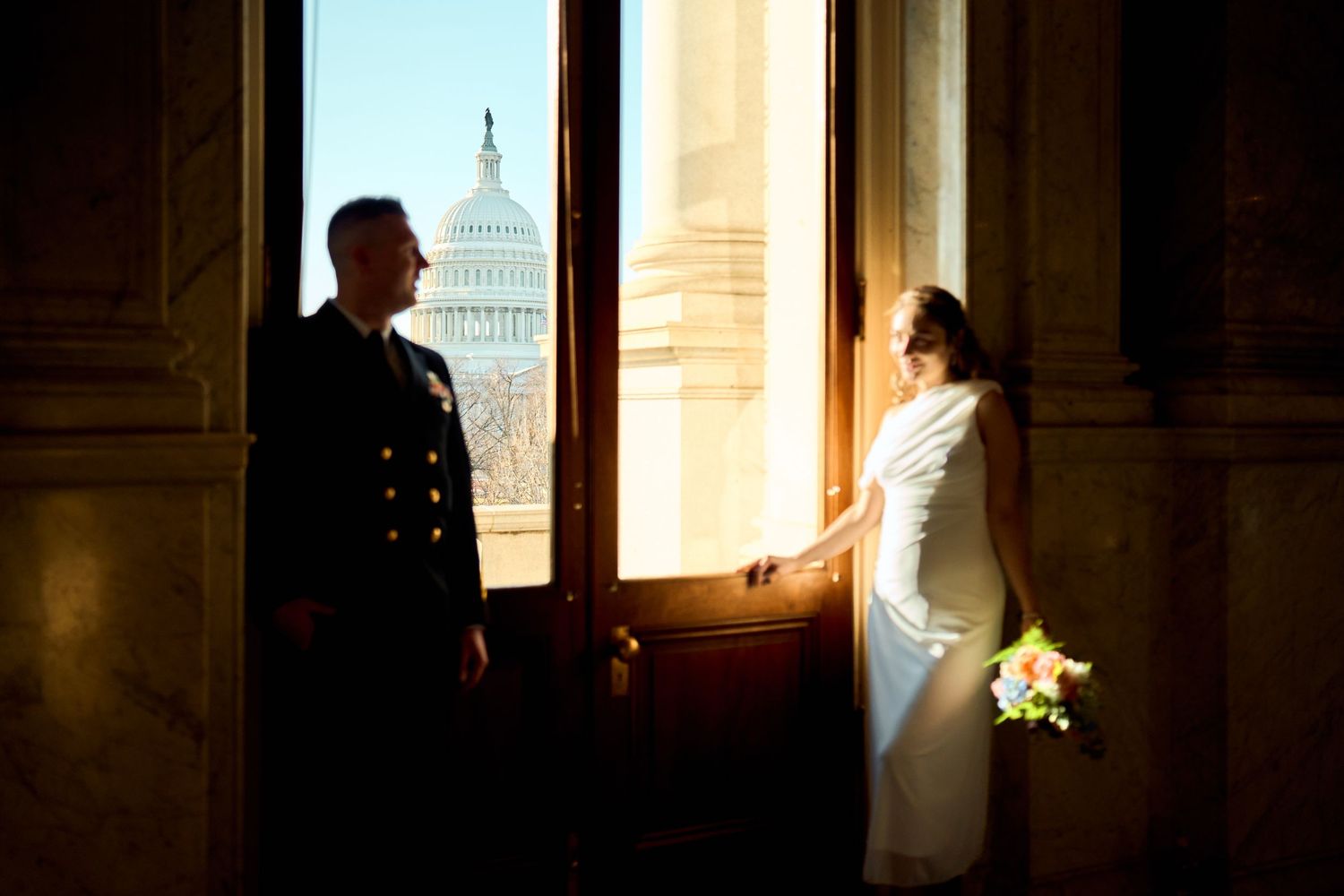 Military groom and bride in white gown holding bouquet pose by open door with US Capitol Building backdrop.