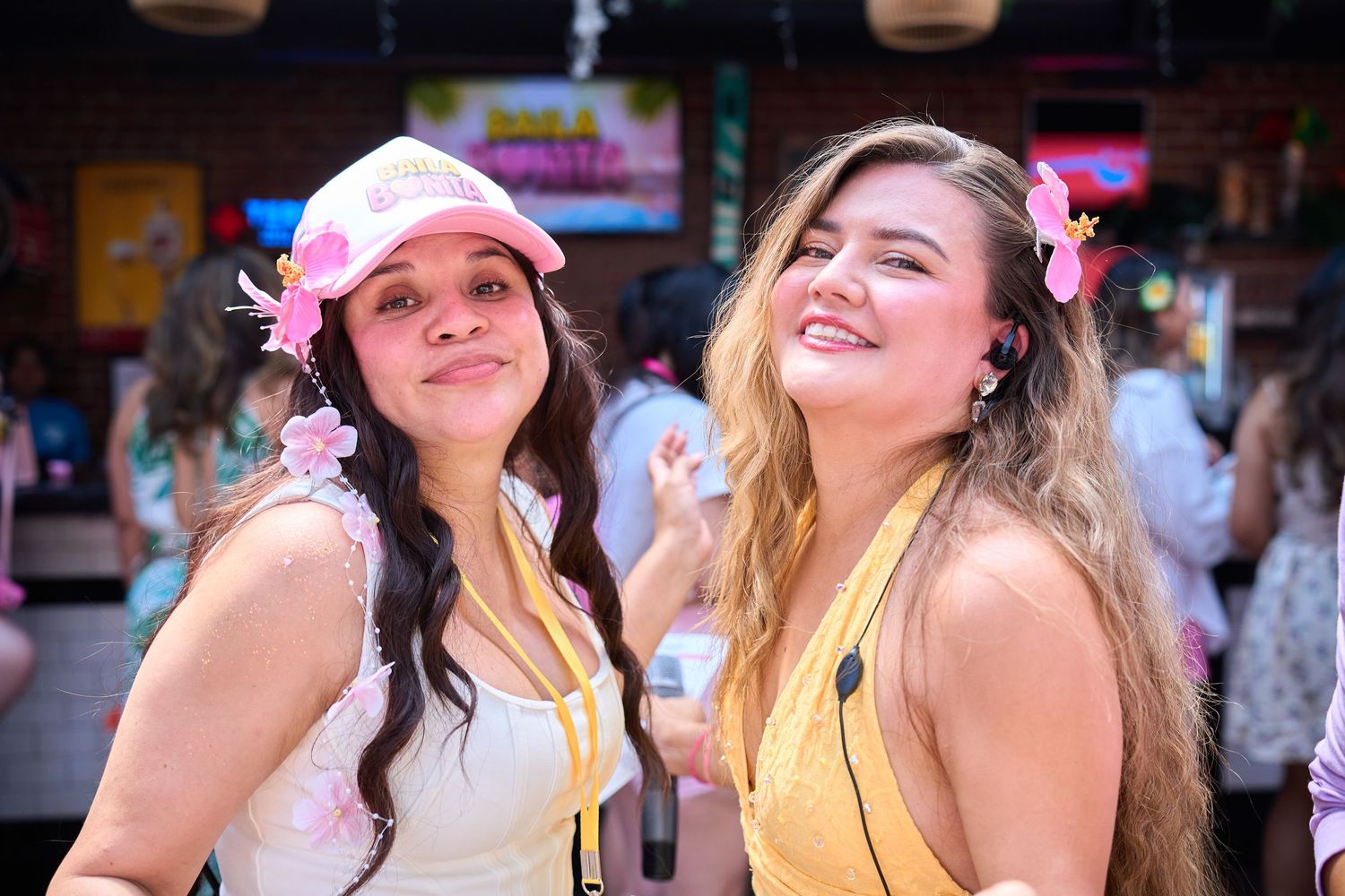 Two smiling women wearing pink floral accessories pose together at a colorful outdoor bar or festival.