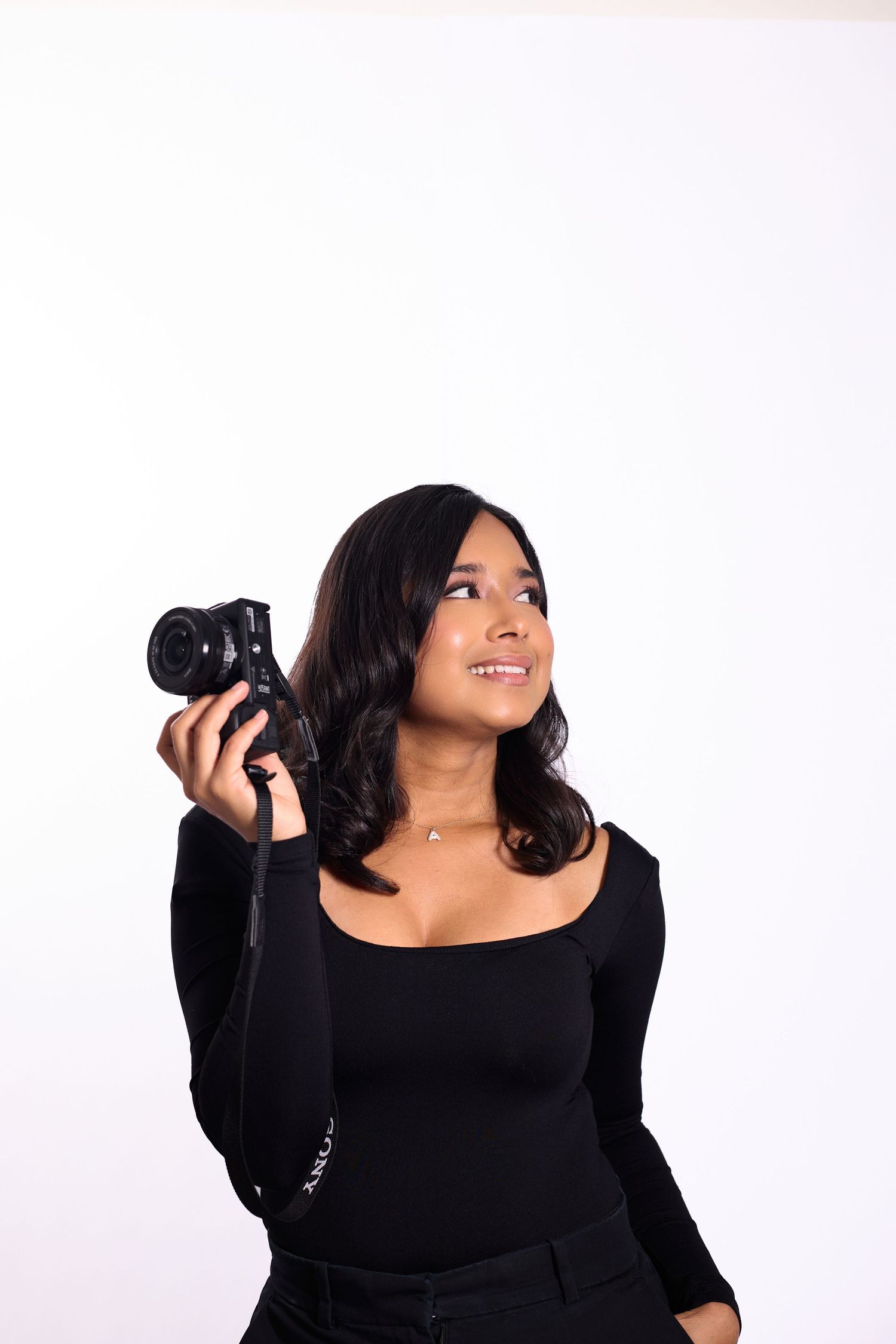 Photographer in black outfit holds camera while posing playfully against white backdrop.