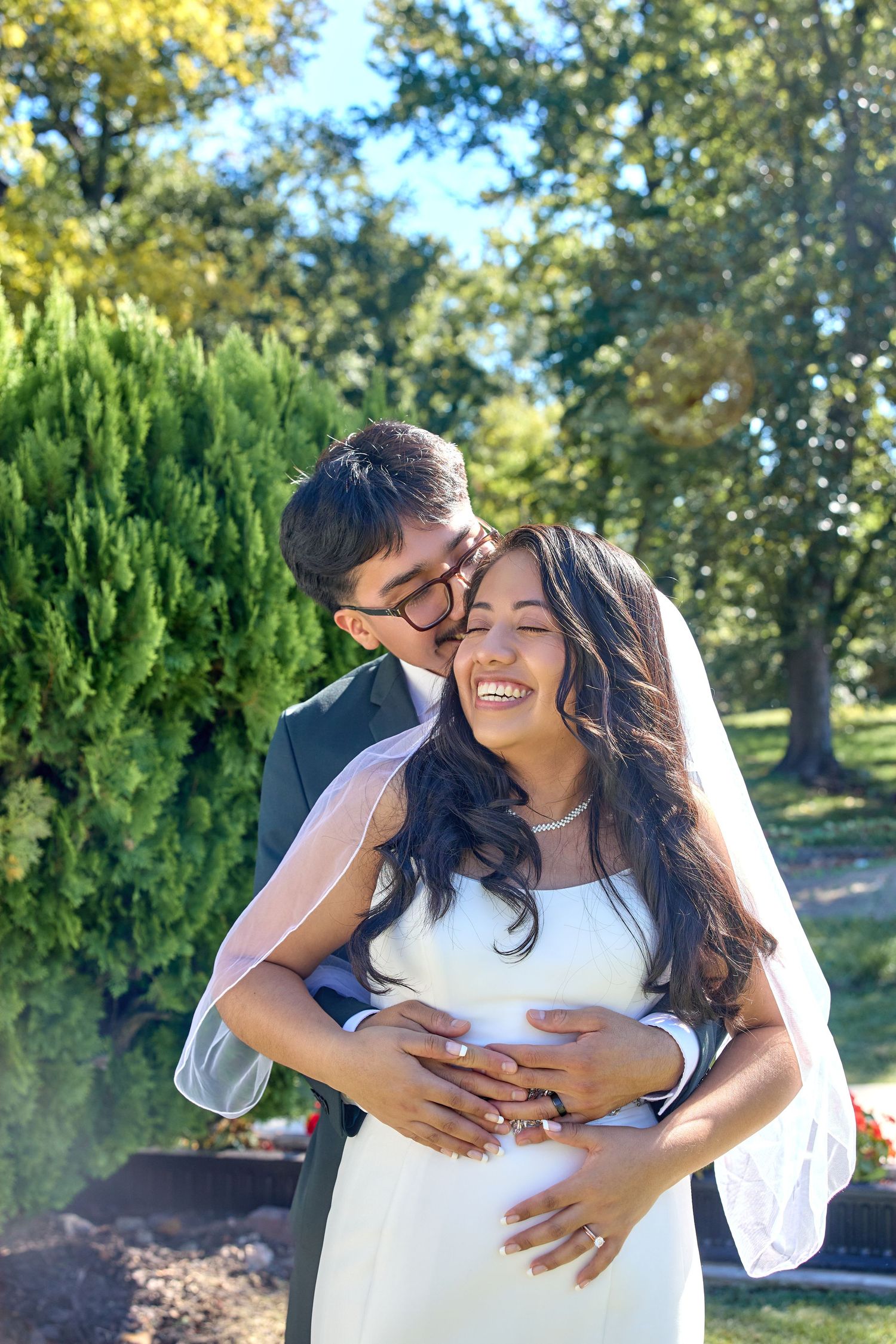 A joyful embrace captured in a sunny garden setting with evergreen trees in the background.