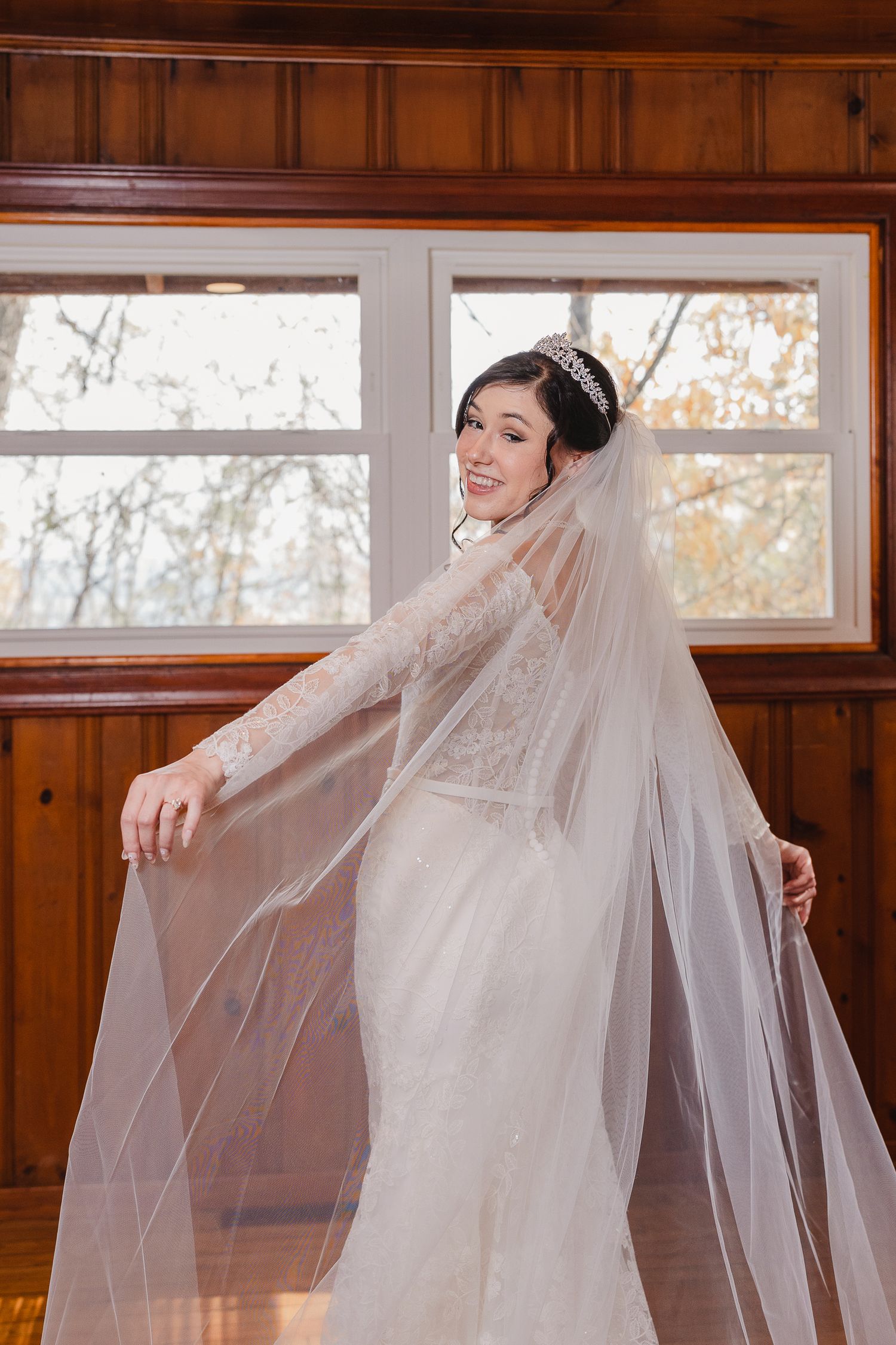 A bride in a lace wedding gown with long veil poses by wooden paneled windows in natural light.