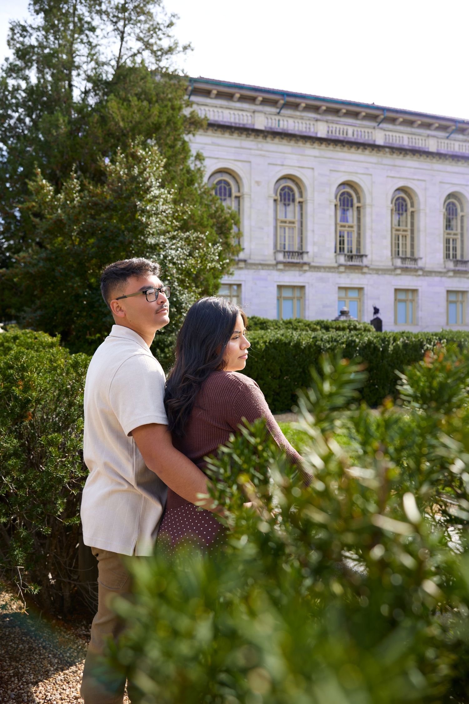 A couple embraces in front of a grand neoclassical building with arched windows and stone facade.