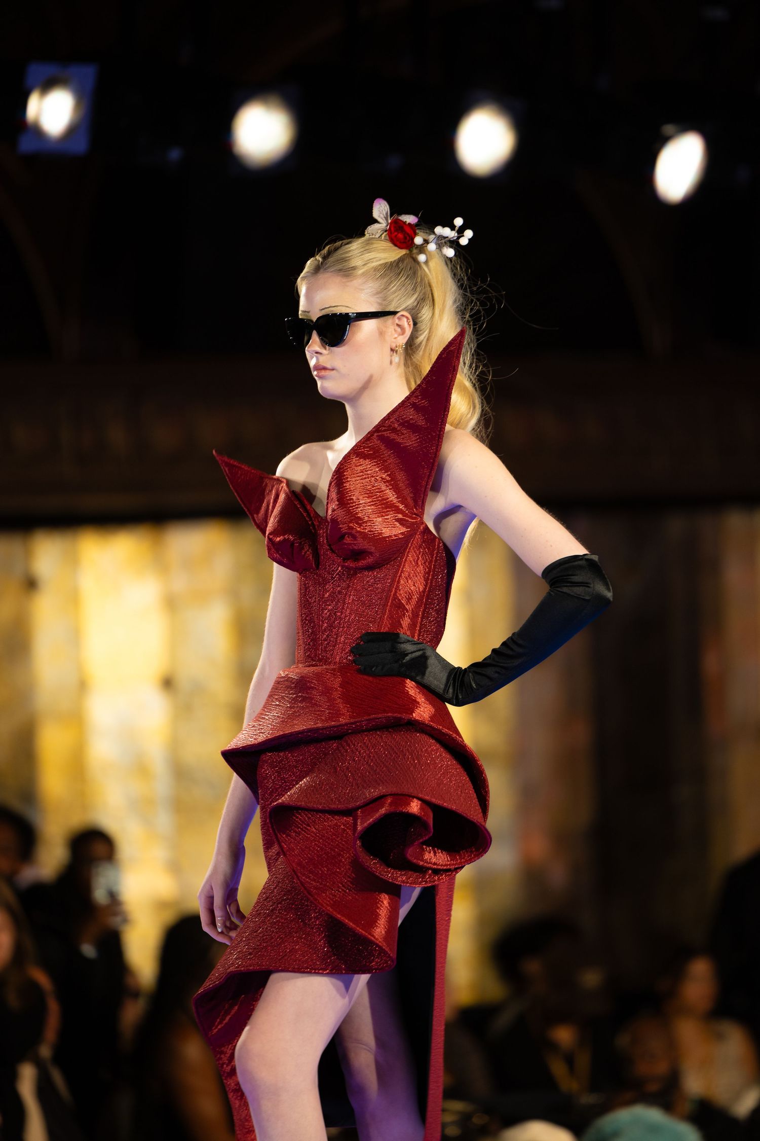 Fashion model wearing red sculptural dress with ruffles, black gloves and sunglasses on runway.
