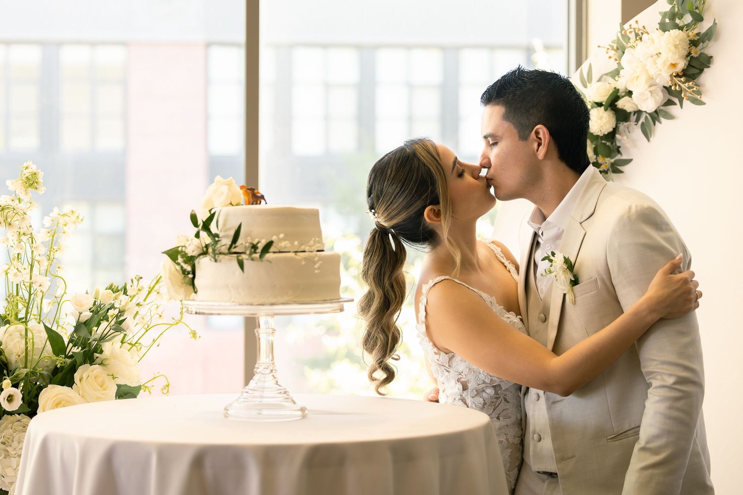 A romantic couple shares a kiss beside their wedding cake at an elegant reception decorated with white flowers.