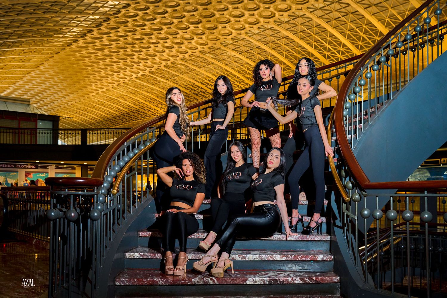 Group of women in black outfits pose on ornate staircase in golden-ceilinged venue.