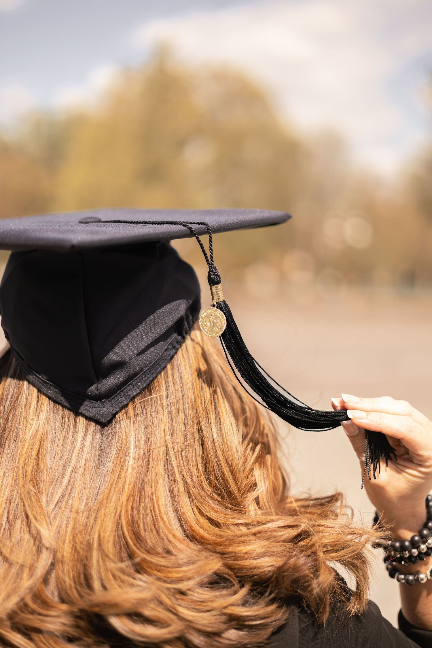 A graduation cap with black tassel being adjusted on blonde wavy hair against a blurred outdoor background.