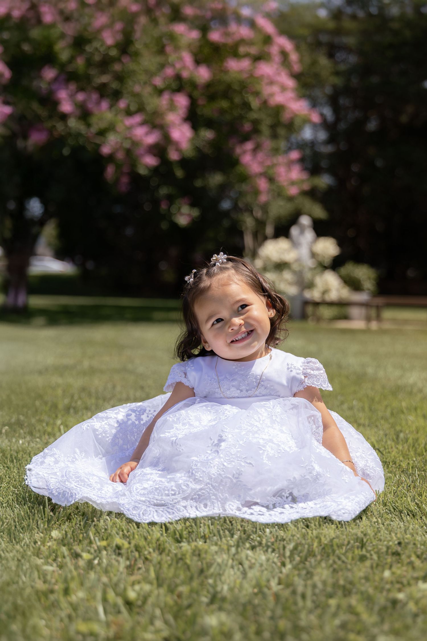 Small child in white lace dress sitting on grass with pink flowering trees in background.