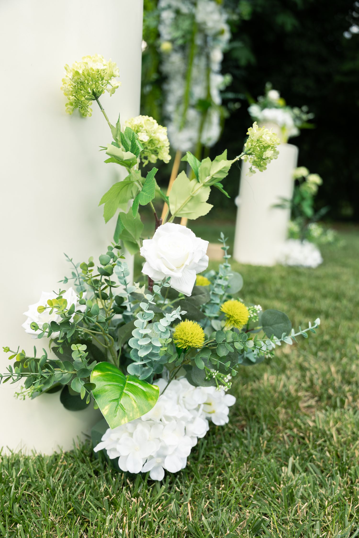 White floral arrangements with eucalyptus greenery line a grassy pathway creating an elegant outdoor decor display.