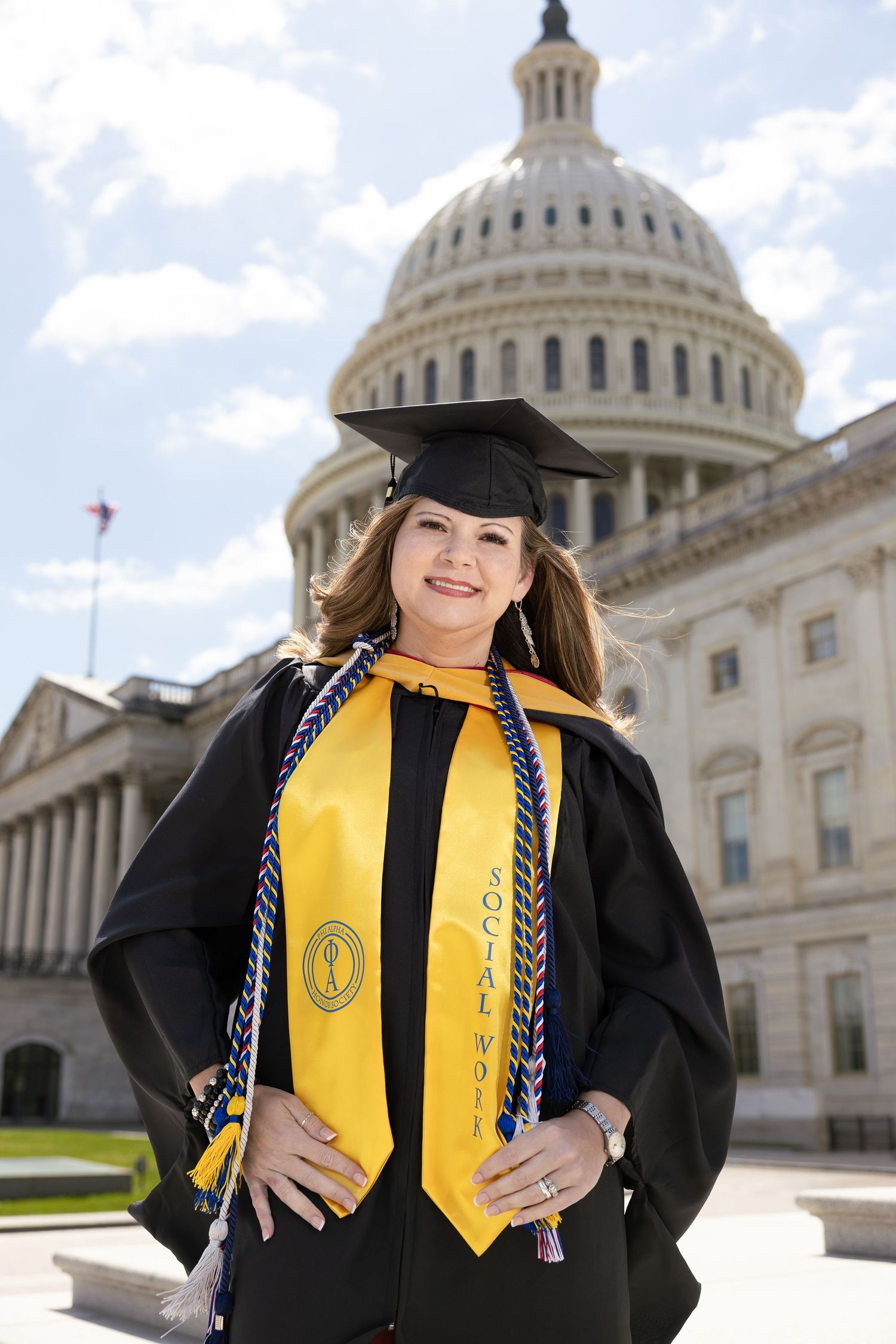 Graduate in cap and gown with yellow stole stands smiling in front of US Capitol building dome on sunny day.