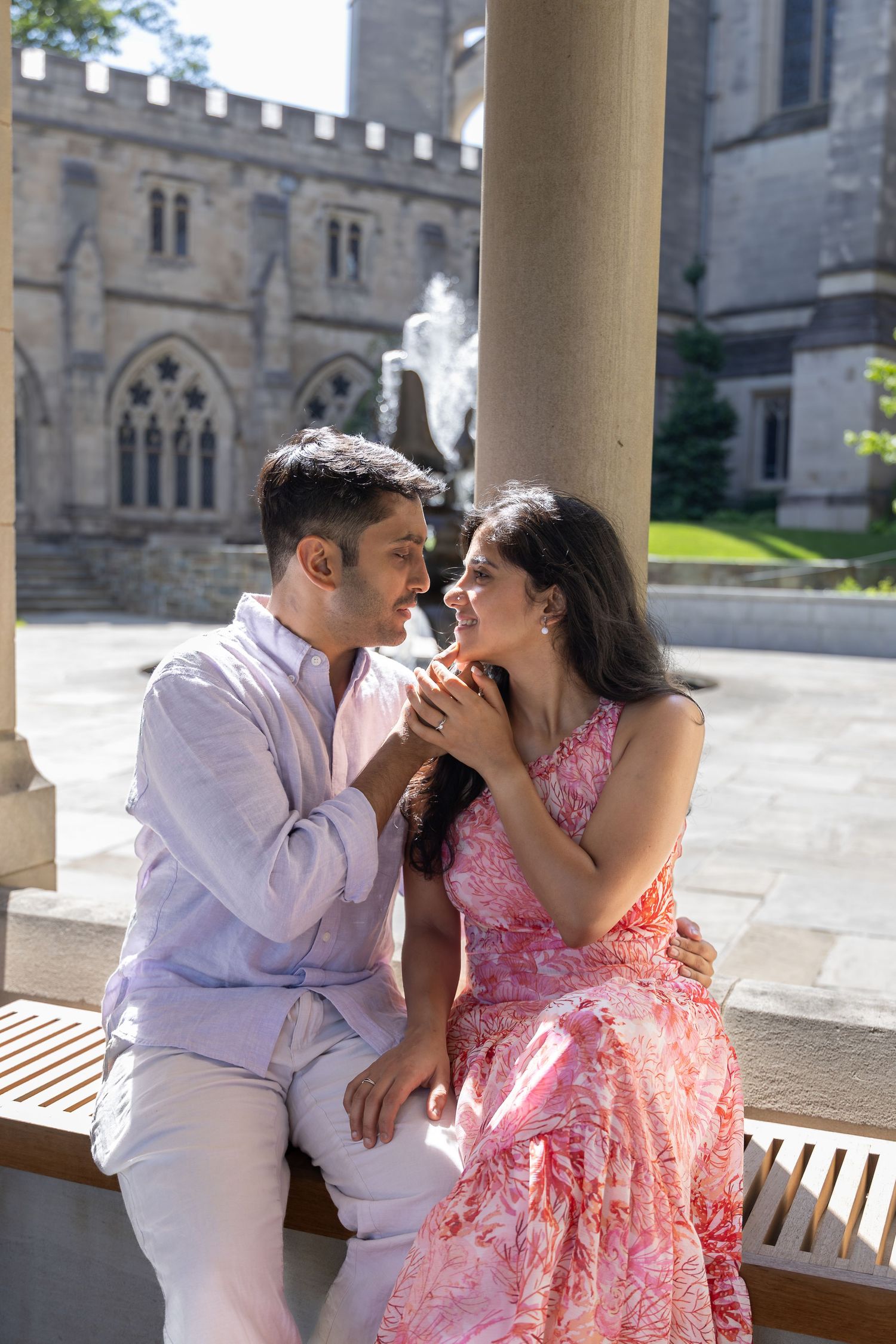 Couple in pink dress and white shirt share romantic moment on a bench with gothic church architecture in background.