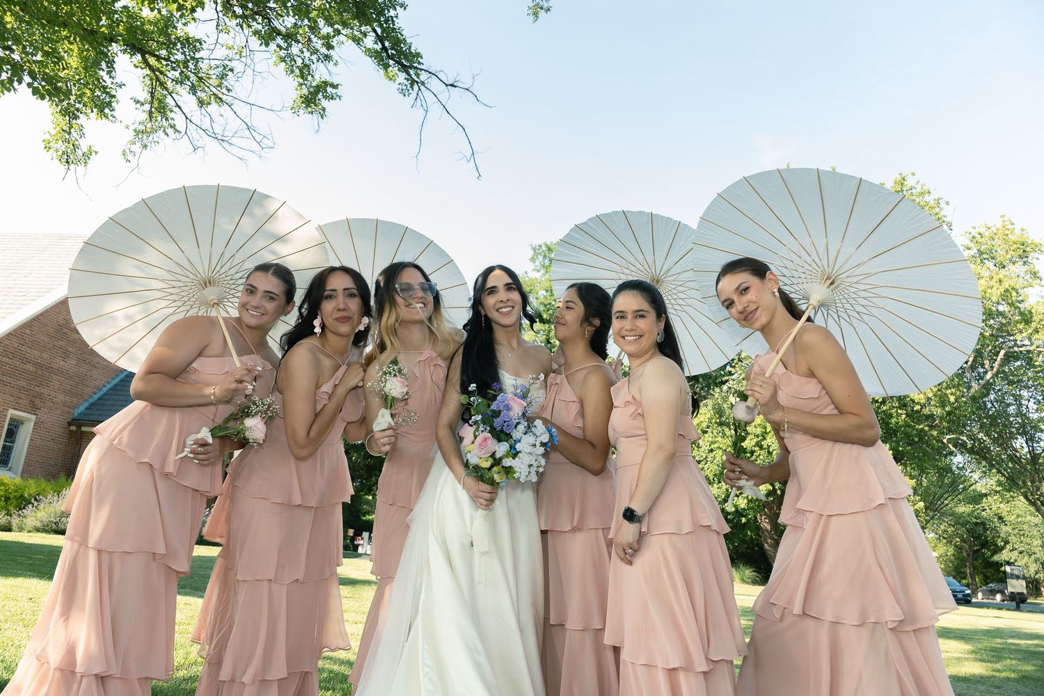 Wedding party in blush pink dresses posing together with white parasols and bouquets outdoors on a sunny day.