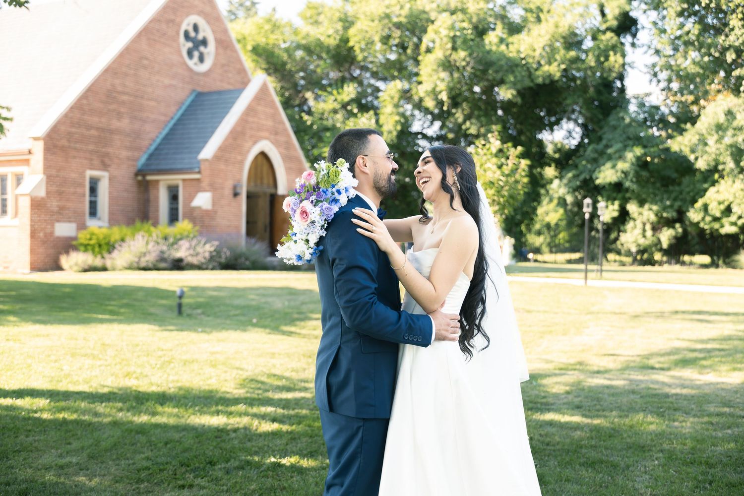 A couple in elegant wedding attire walks outdoors with a church building in the background.