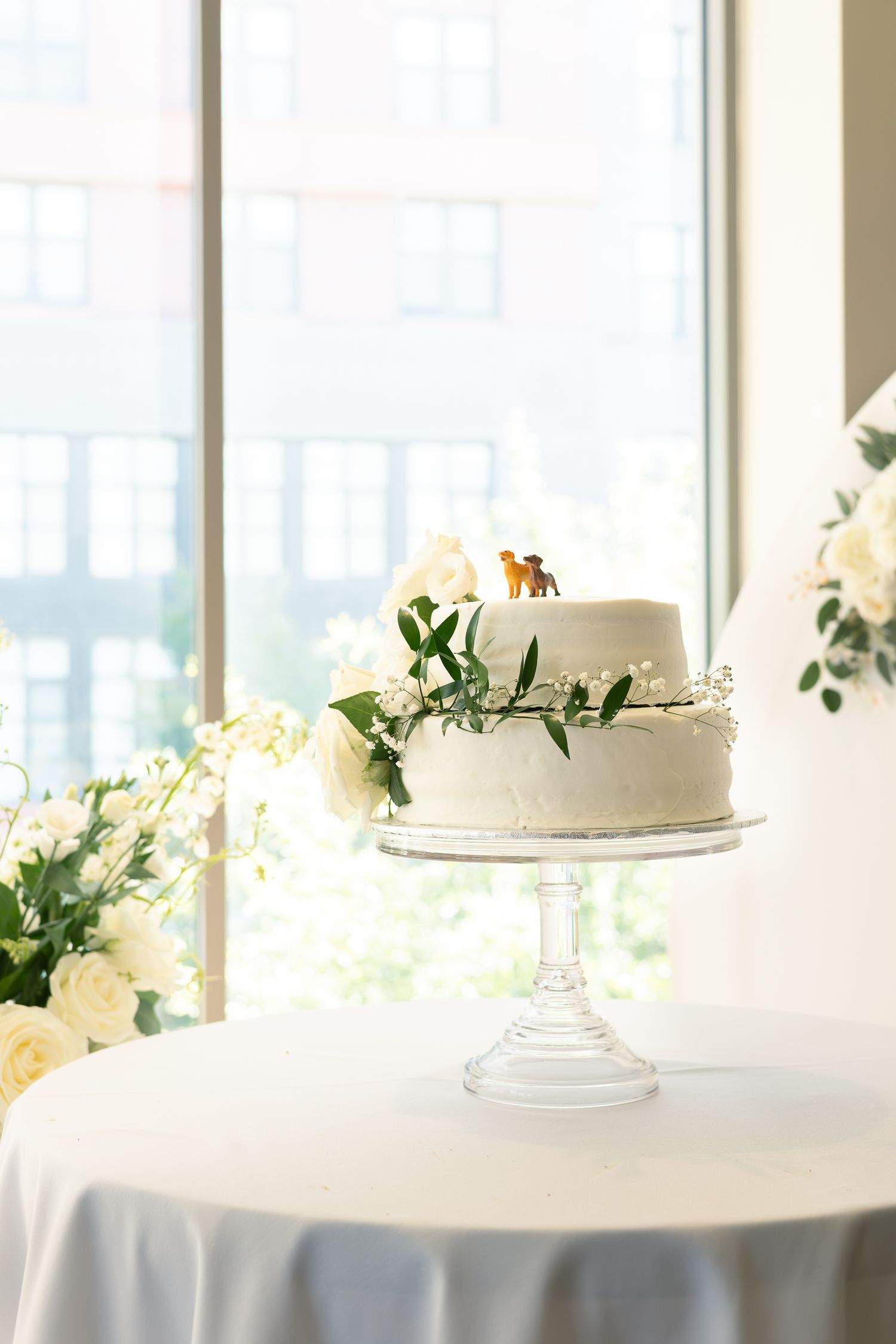 White wedding cakes displayed on glass stands with floral decorations on elegant white tablecloths in a bright venue.